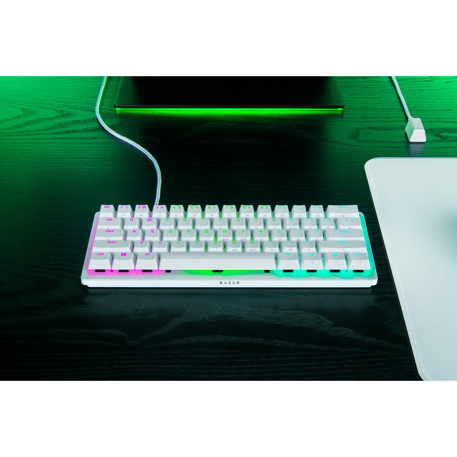 A white Razer keyboard with colorful backlit keys sits on a black desk, near a monitor and a white mousepad. The Razer logo is visible.
