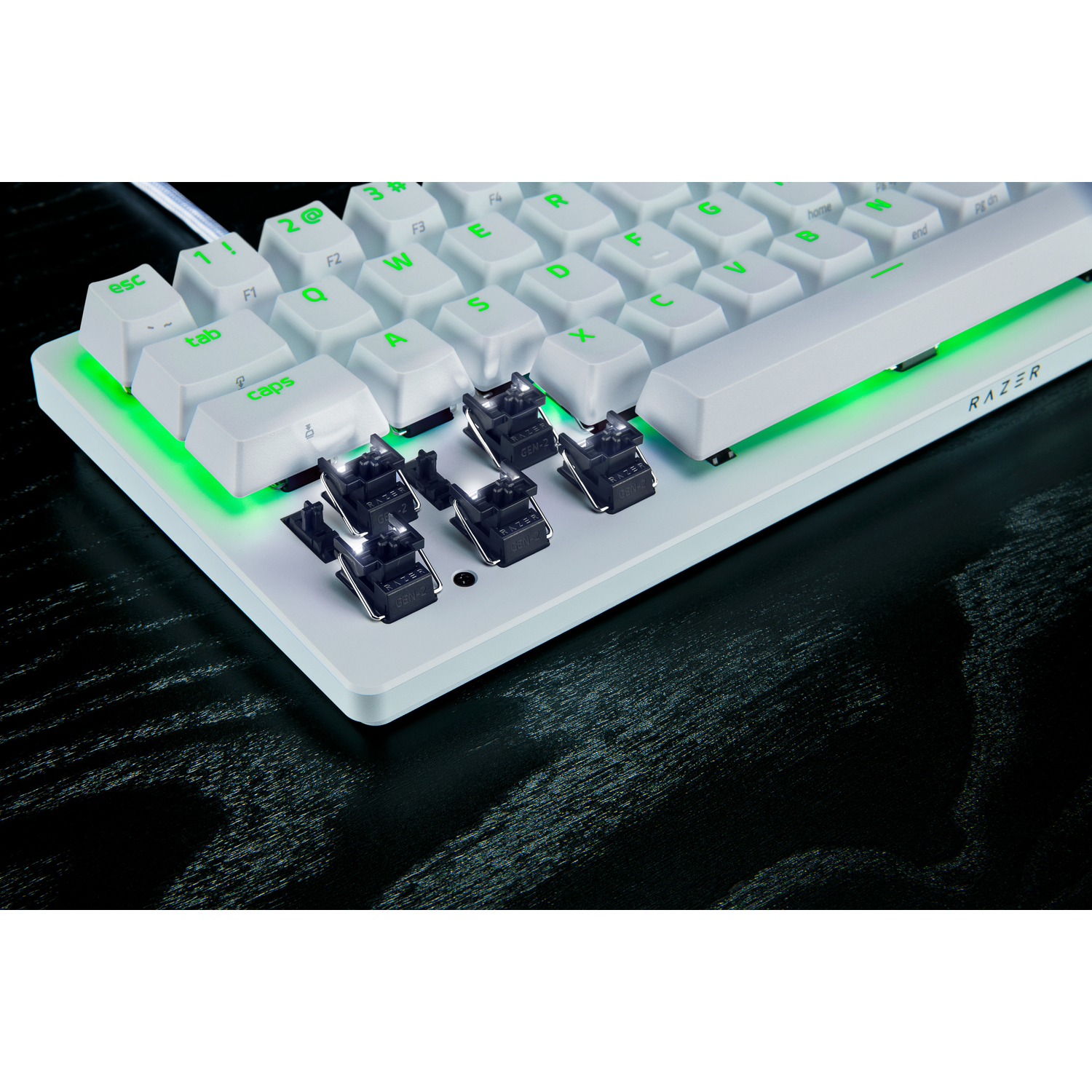 A white Razer keyboard with green illuminated keys. Some keys are removed to show the switches. The keyboard is on a black surface.