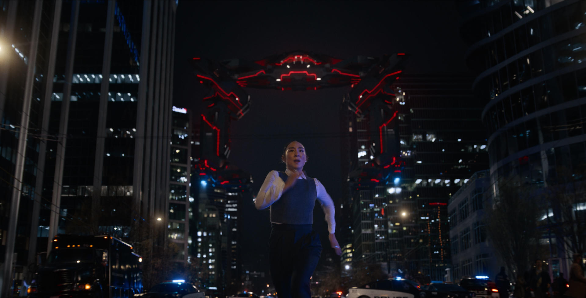 Eine Frau rennt auf eine große, rot beleuchtete Struktur in einer Stadt bei Nacht zu. Wolkenkratzer und Polizeiautos sind im Hintergrund zu sehen.