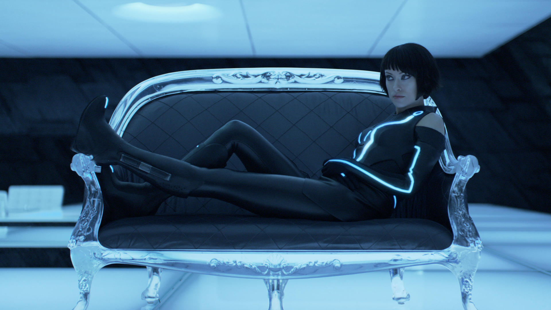 Eine Frau in einem schwarzen Anzug mit blauen Neonlinien liegt in futuristischer Umgebung auf einer silbernen Couch.