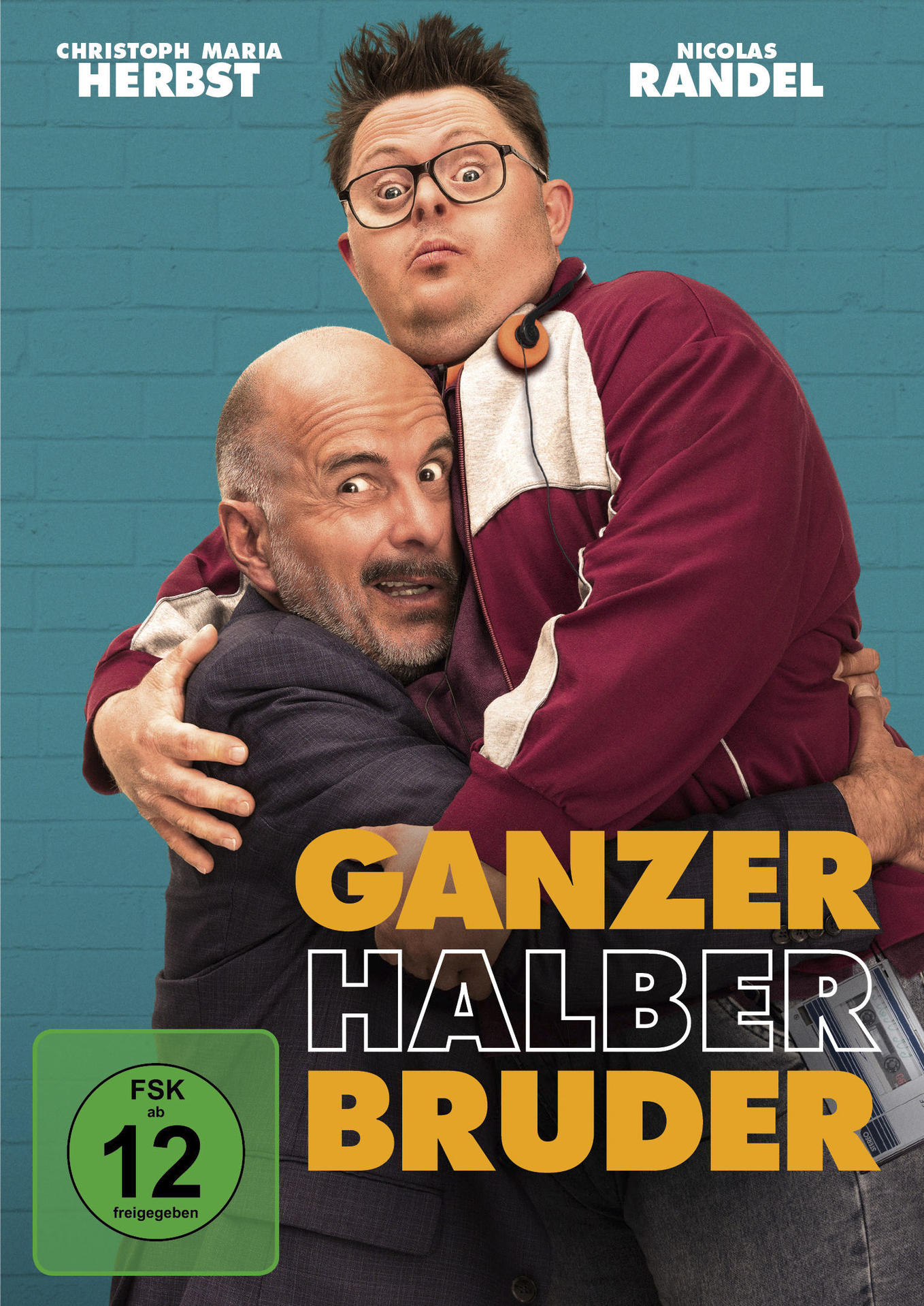 Poster für den Film „Ganze halbe Bruder“. Zwei Männer umarmen sich, einer trägt Brille und eine rote Jacke. Der Hintergrund ist eine blaue Ziegelmauer.