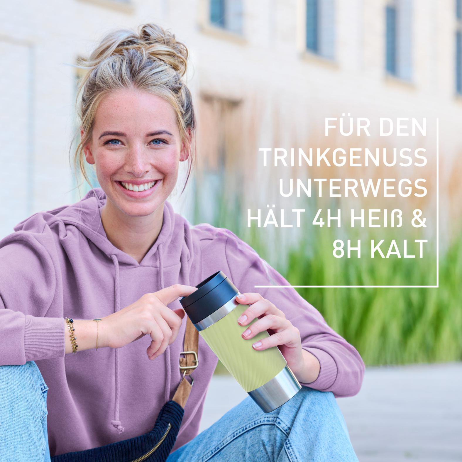 Eine lächelnde Frau in einem lila Kapuzenpullover hält eine grüne Thermoskanne. Text: Genießen Sie Getränke unterwegs; hält Getränke 4h heiß und 8h kalt.