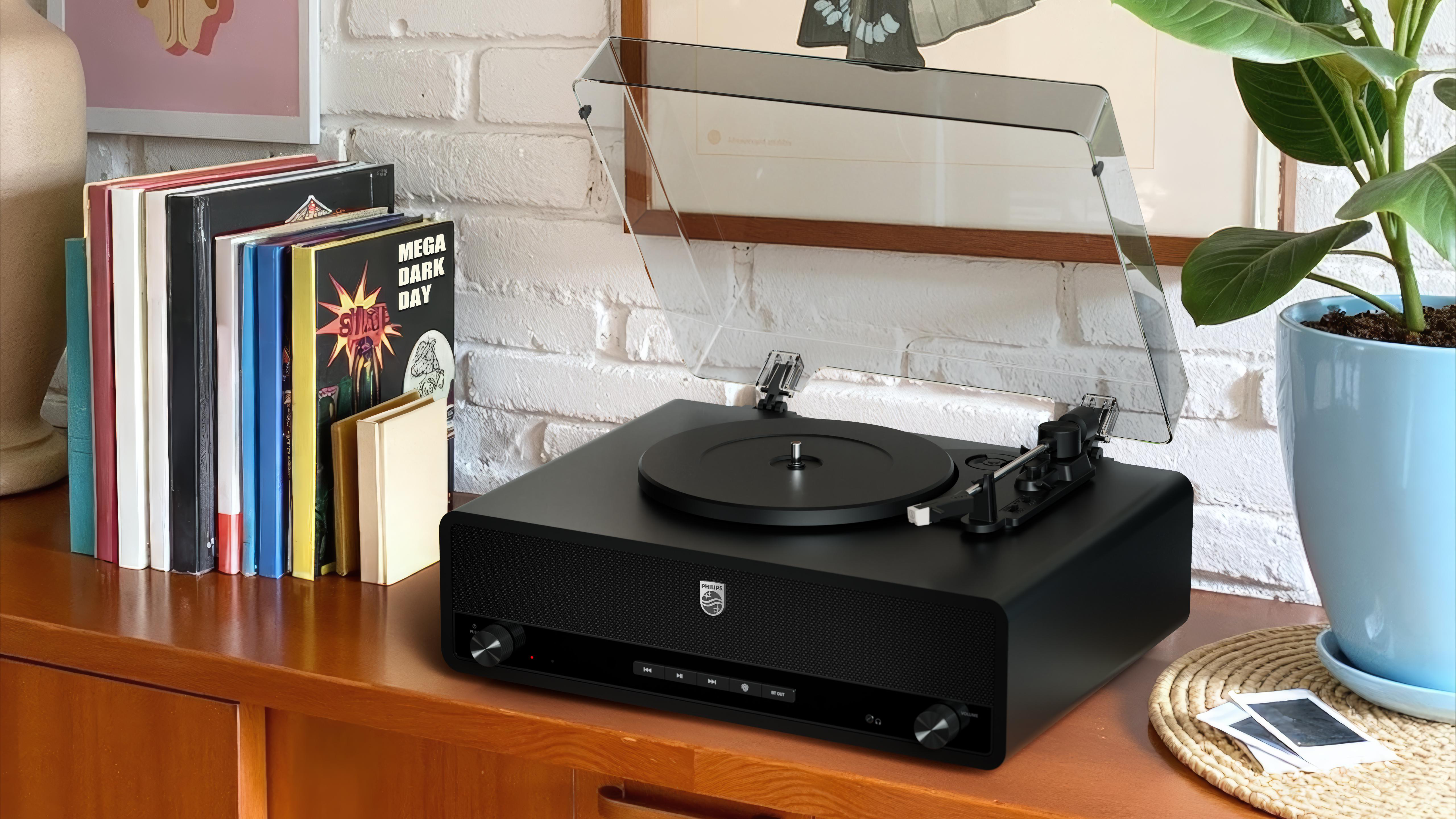 Schwarzer Plattenspieler auf Holzoberfläche mit Schallplatten, Pflanze und gerahmter Kunst im Hintergrund.