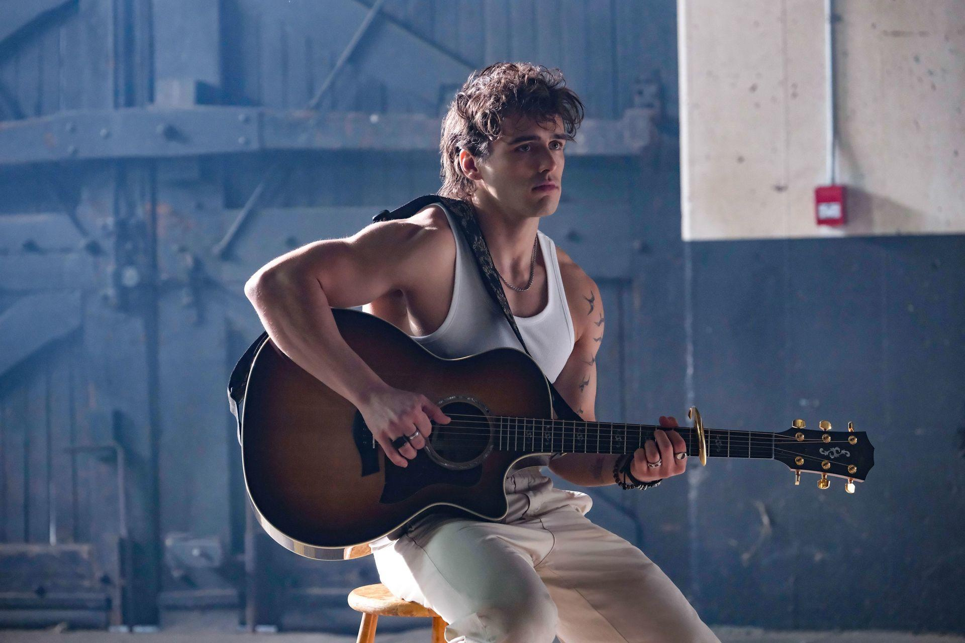 Ein Mann in einem weißen Tanktop spielt eine Akustikgitarre. Er sitzt auf einem Hocker in einer Lagerhalle und wirkt nachdenklich.