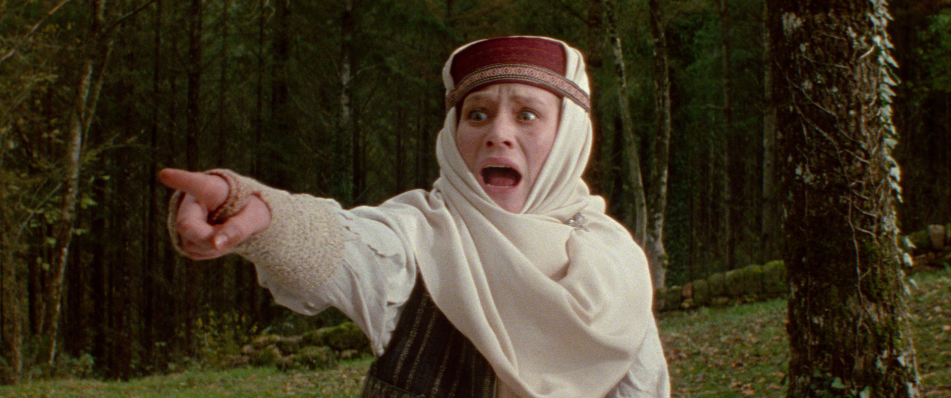 Eine Frau in traditioneller Kleidung, die mit dem Finger zeigt und schreit, mit aufgerissenen Augen, steht in einem Wald. Bäume und Gras im Hintergrund.