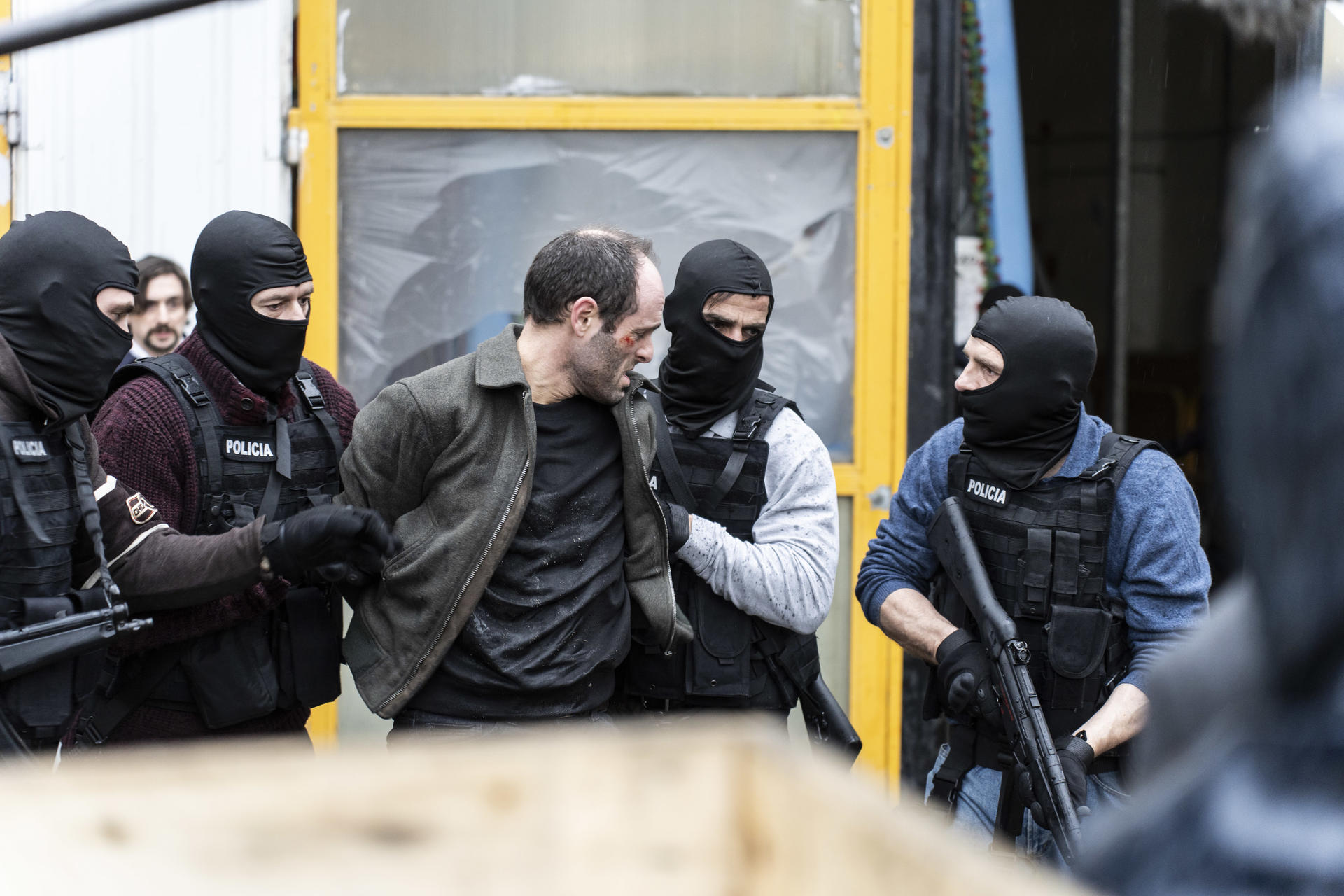 Ein Mann wird von Polizisten in Sturmhauben festgenommen. Die Beamten tragen Westen. Der Mann hat eine Wunde. Hintergrund: ein gelbes Gebäude.