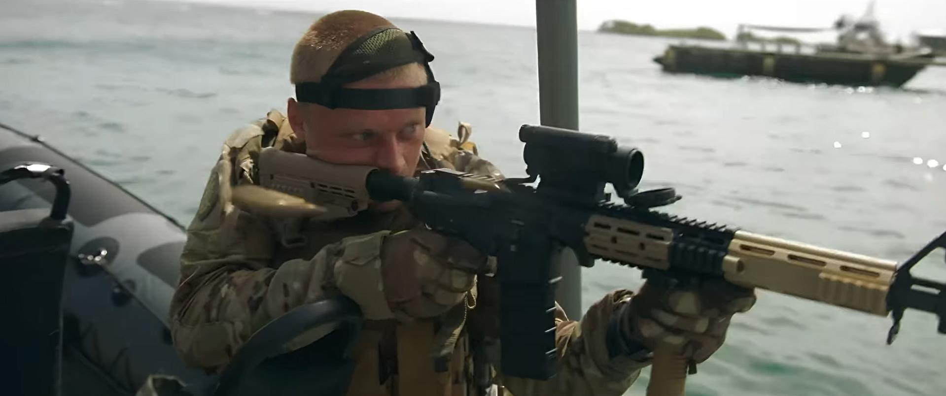 Ein Soldat zielt mit einem Gewehr von einem Boot aus auf Wasser. Er trägt Tarnung und ein Headset. Ein zweites Boot ist im Hintergrund.