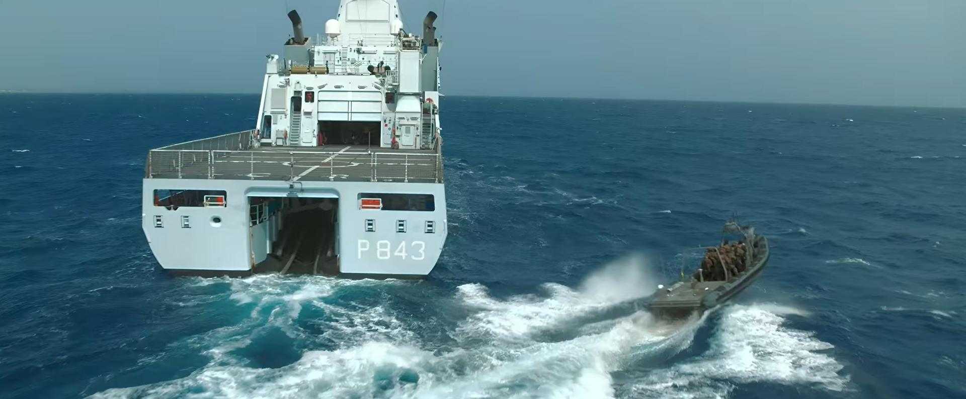 Ein großes Marineschiff mit der Nummer P843 und ein kleines Boot im Wasser. Das Meer ist blau und der Himmel bedeckt.