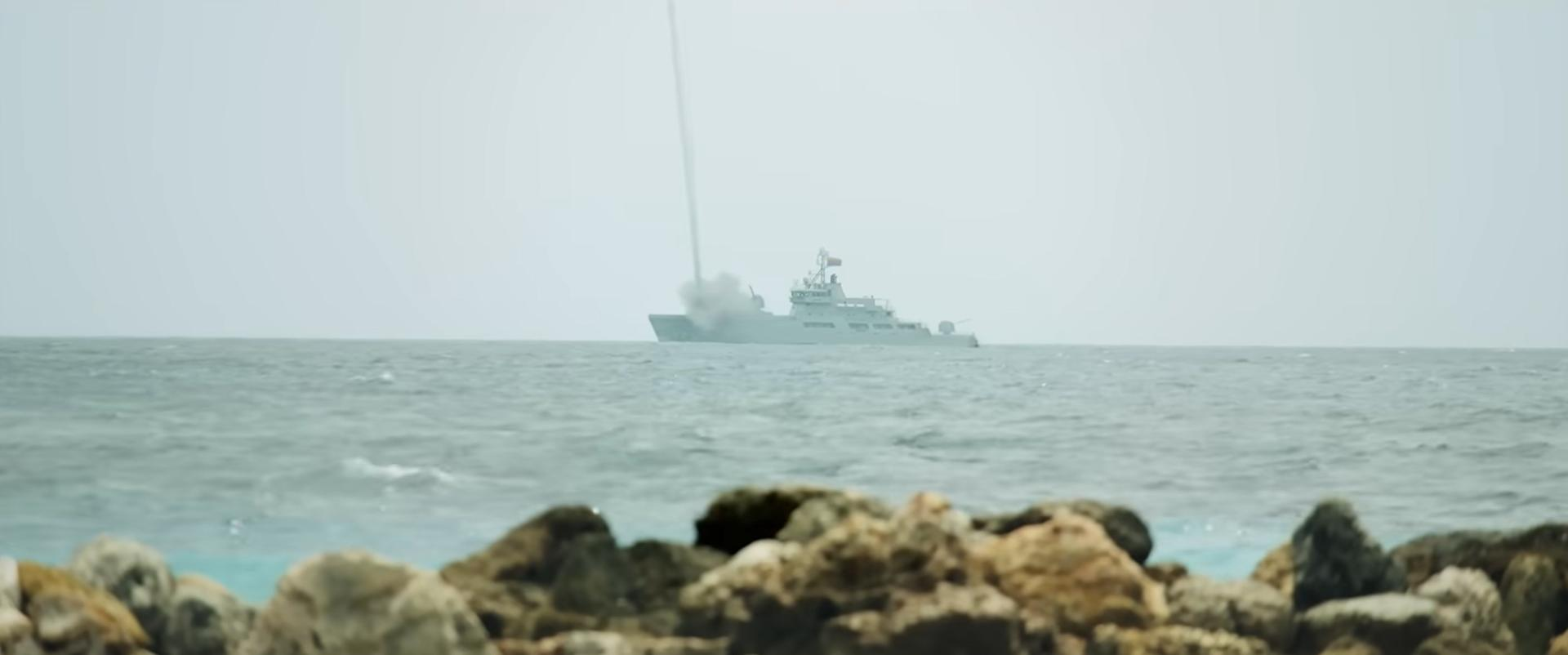 Ein Kriegsschiff feuert eine Rakete in den Himmel. Im Vordergrund befindet sich das Meer, im Hintergrund Felsen und das Schiff.