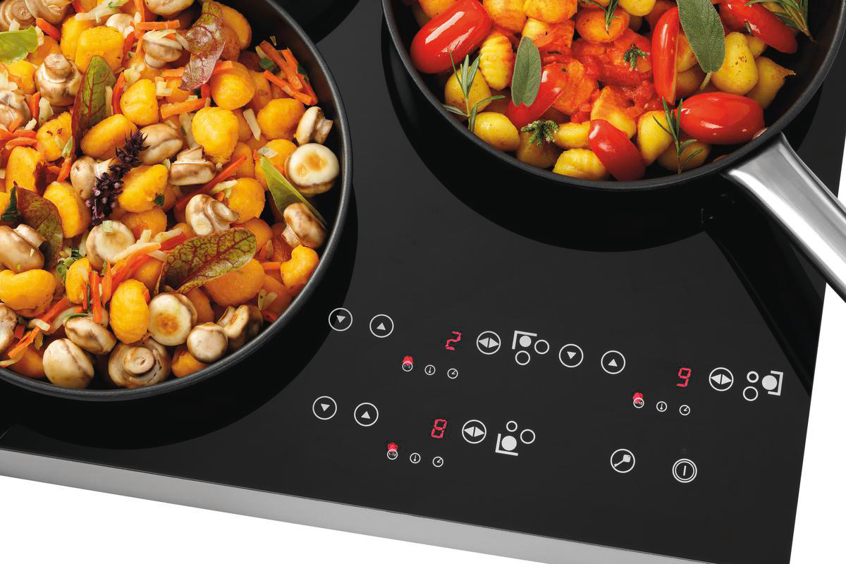 Zwei Pfannen auf einem schwarzen Herd. Eine Pfanne hat Pilze und Karotten. Die andere hat Tomaten und Kräuter. Herdsteuerungen sind sichtbar.