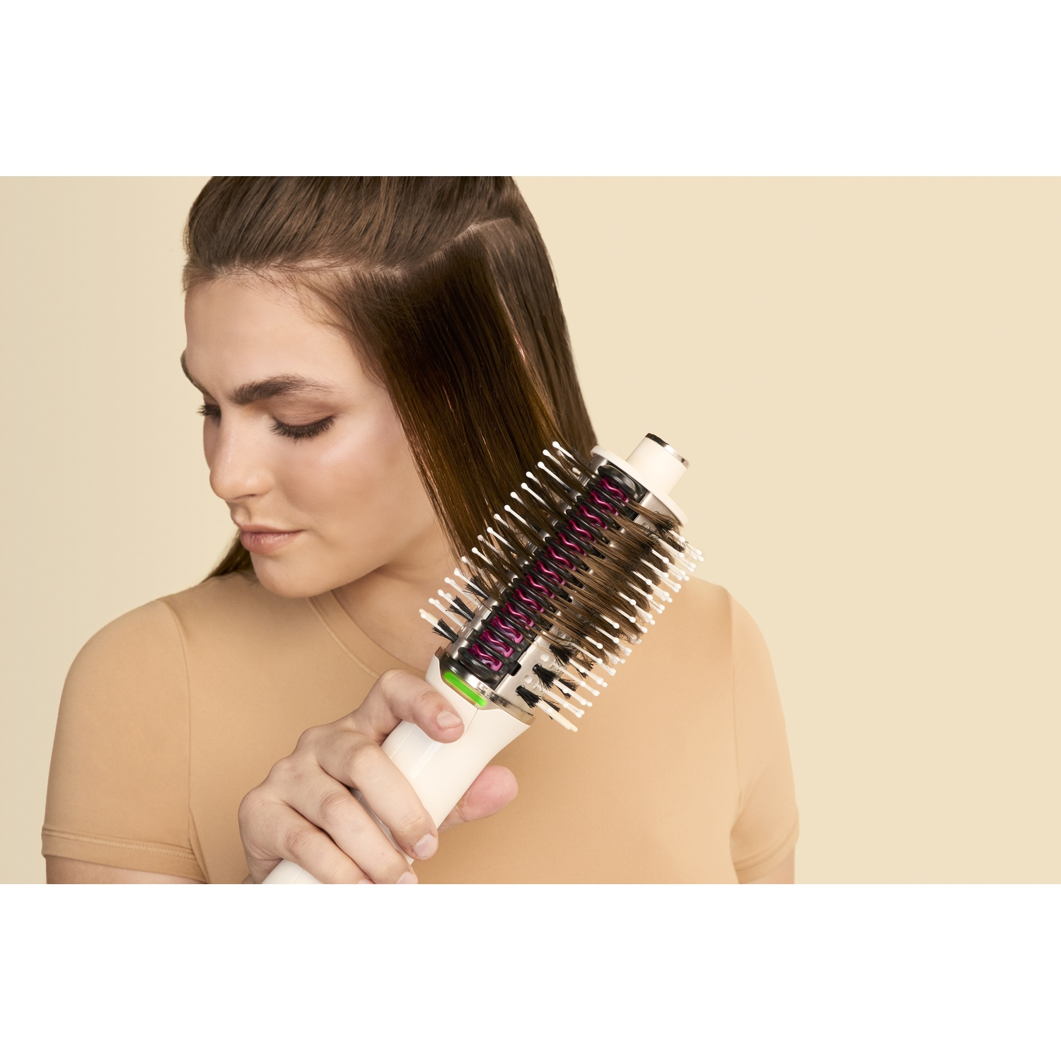 Een vrouw gebruikt een haarborstel op haar bruine haar. Ze draagt een beige top, en de achtergrond is ook beige.