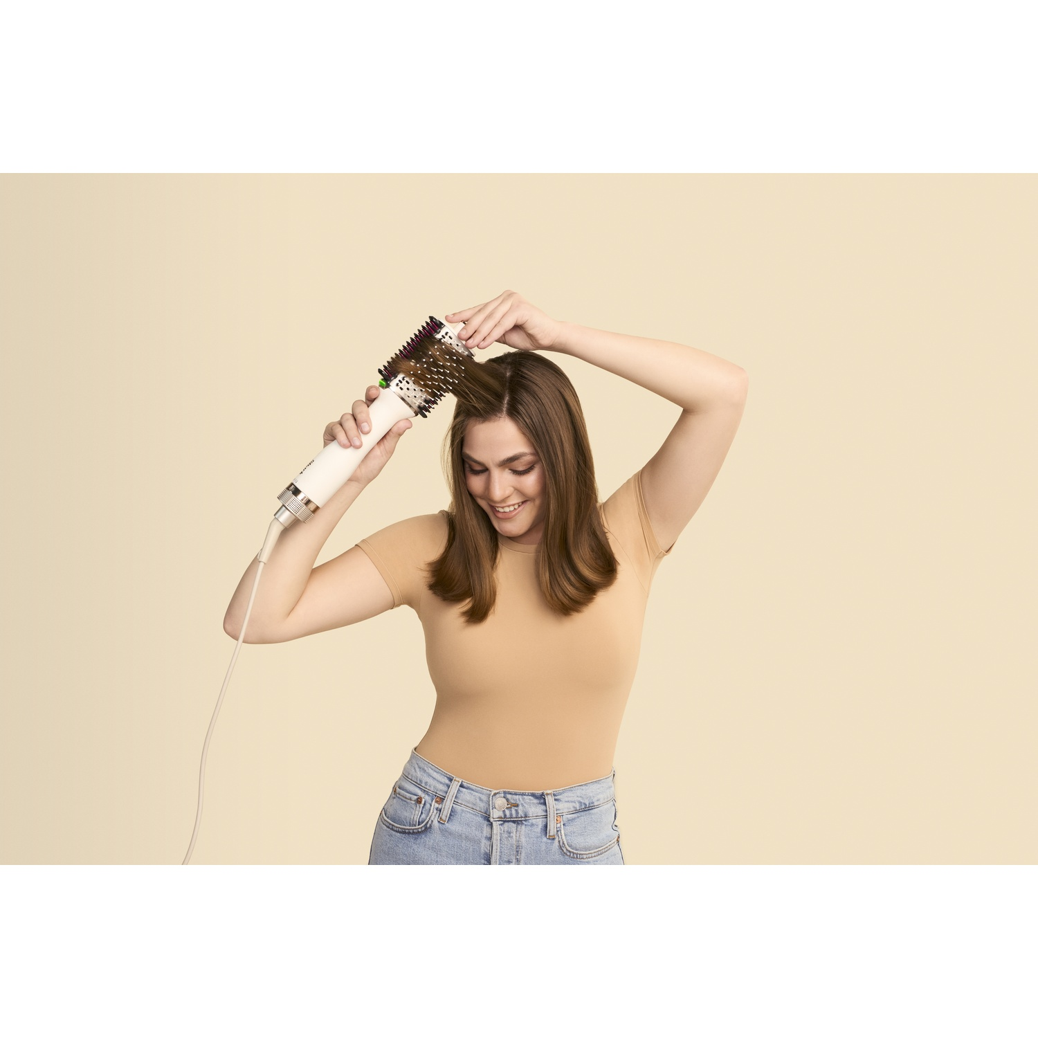 Een vrouw gebruikt een haarborstel. Ze draagt een beige shirt en een spijkerbroek en glimlacht. De borstel is wit en bruin.