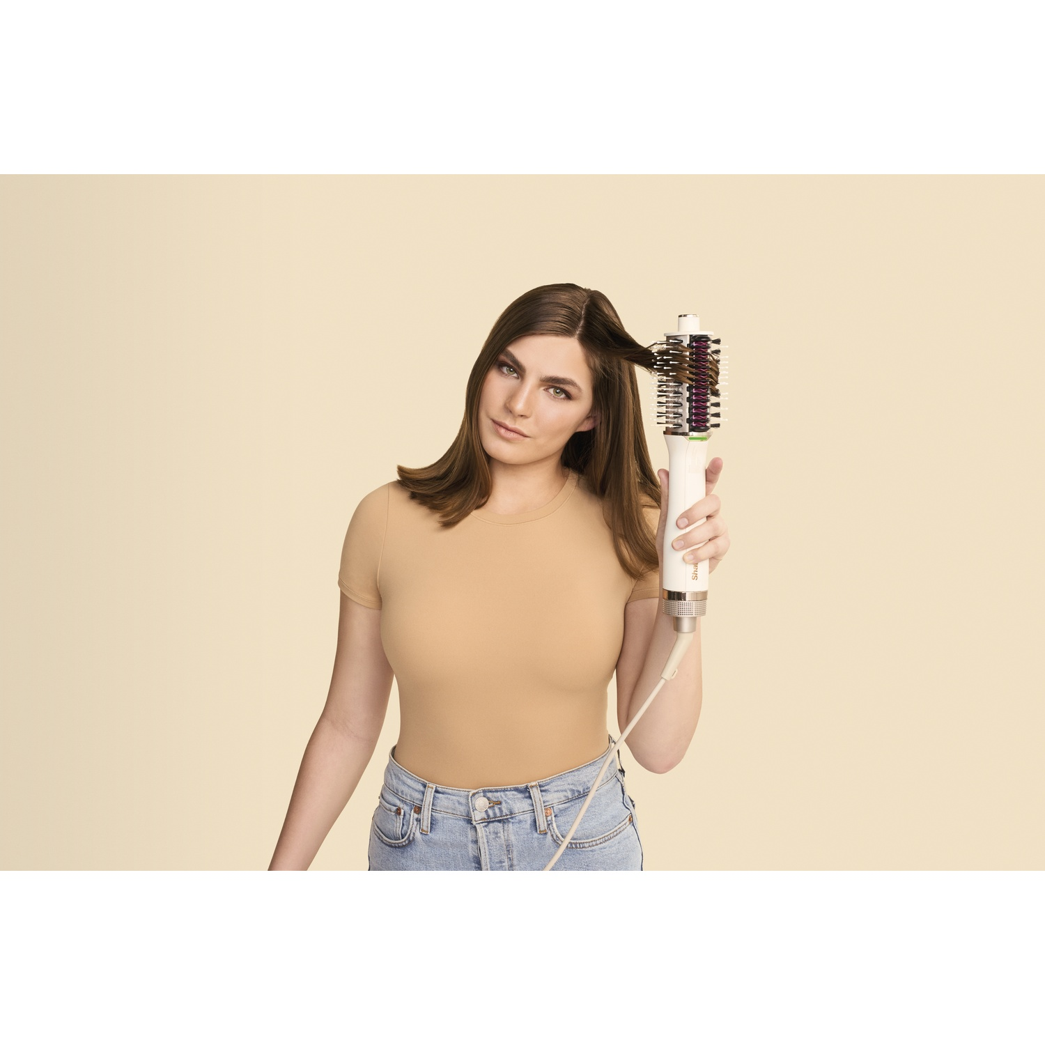 Een vrouw houdt een witte haarstylingtool vast, waarmee ze haar bruine haar stylet. Ze draagt een beige shirt en jeans tegen een lichte achtergrond.