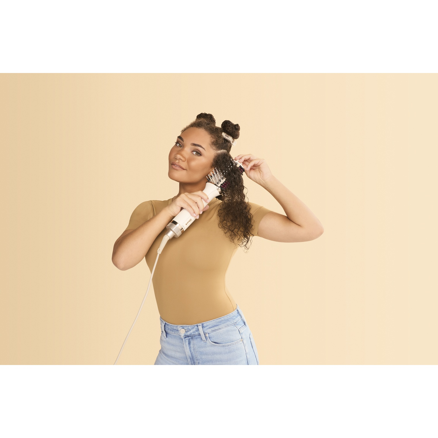 Een vrouw gebruikt een haarstylingtool. Ze draagt een bruin shirt en een spijkerbroek. De achtergrond is lichtbeige.