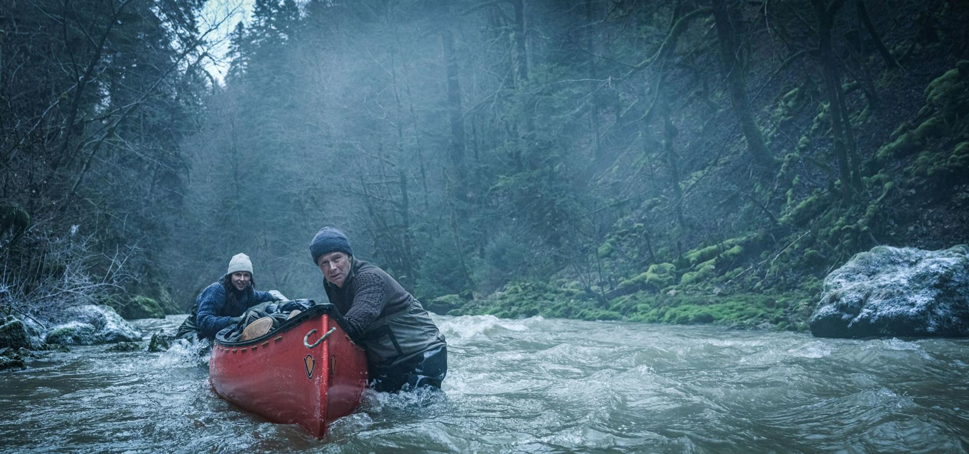 Zwei Personen in einem roten Kanu navigieren einen Fluss. Wald umgibt sie. Der Mann steht im Wasser.