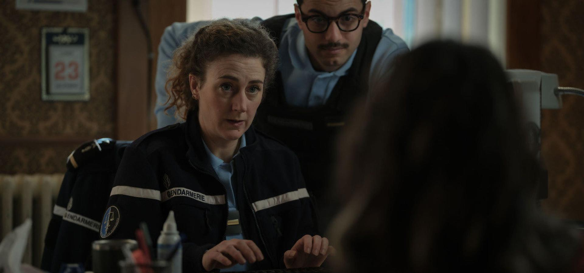 Eine Frau in Gendarmerie-Uniform blickt eine Person an. Ein Mann mit Brille steht hinter ihr. Ein Kalender zeigt das Datum 23.