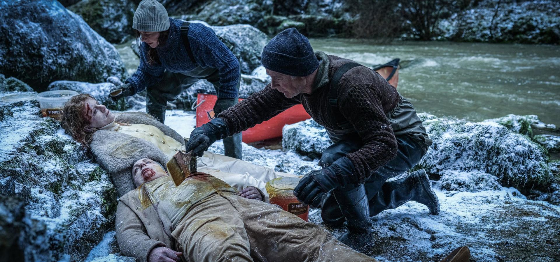 Drei Personen an einem verschneiten Fluss. Einer kniet und trägt einer Person etwas auf die Brust auf. Zwei andere beobachten.