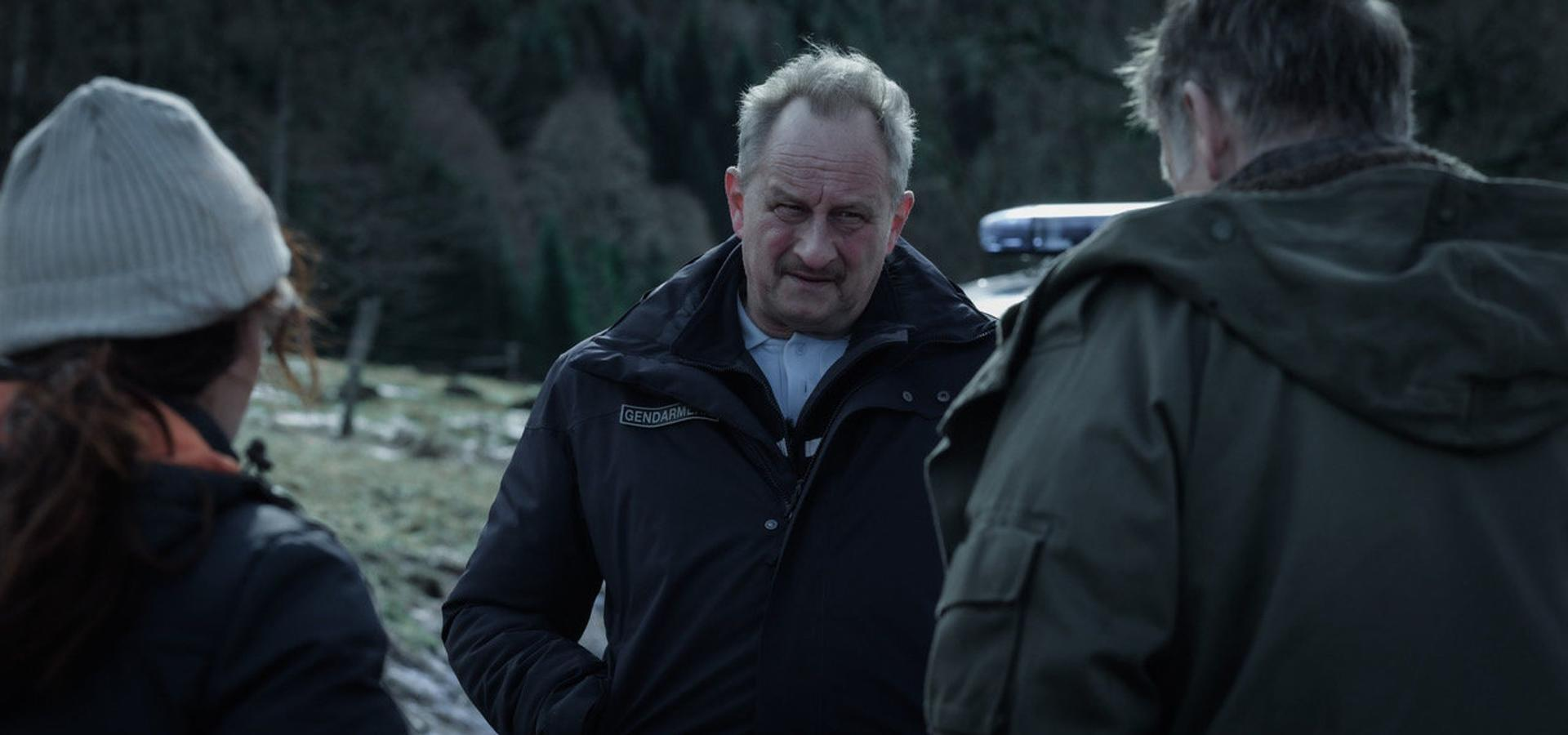 Drei Personen in einer dunklen Szene. Ein Mann in schwarzer Jacke und eine Frau mit Hut stehen einem anderen Mann gegenüber. Wald im Hintergrund.