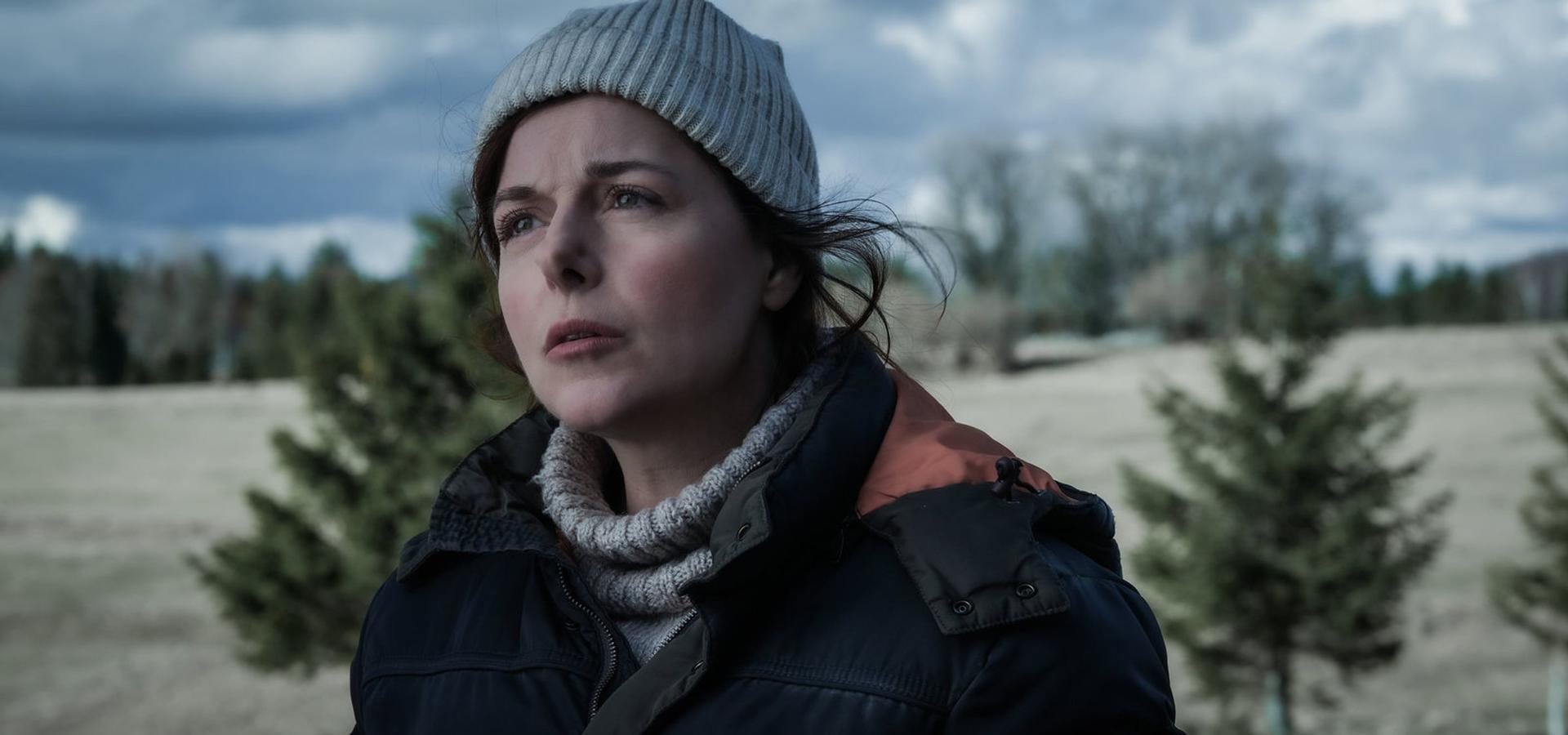 Eine Frau mit grauem Hut und Jacke blickt nach oben. Bäume und ein bewölkter Himmel sind im Hintergrund. Die Szene ist im Freien.