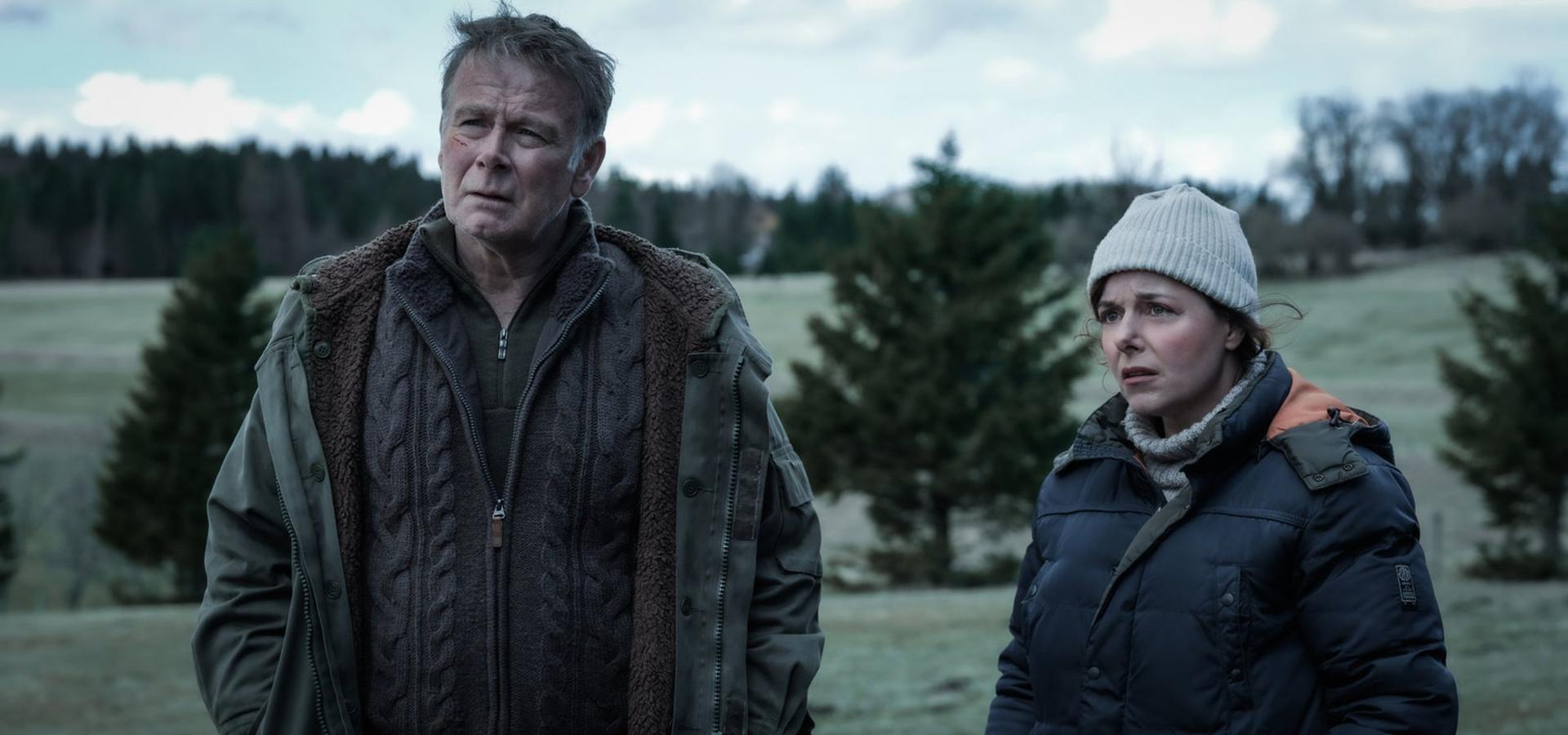 Zwei Personen auf einem Feld. Mann in grüner Jacke, Frau in Mütze. Bäume im Hintergrund. Bewölkter Himmel.
