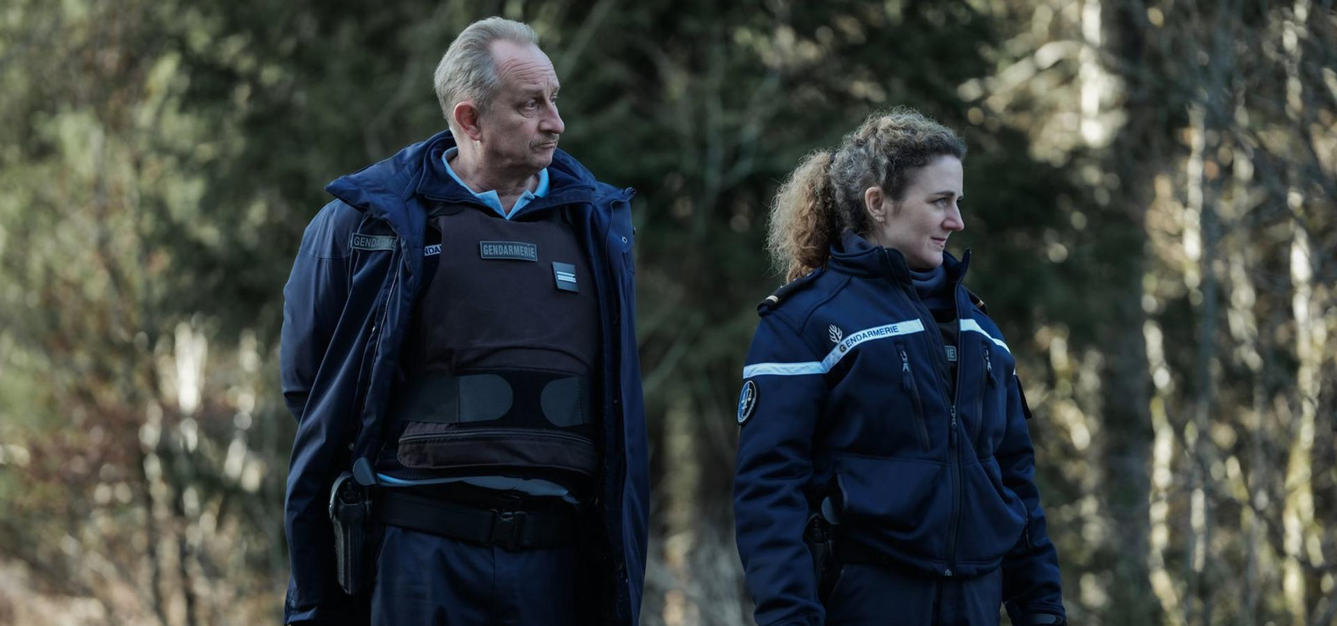 Zwei Gendarmen in blauen Uniformen stehen im Freien. Ein Mann trägt eine kugelsichere Weste. Bäume sind im Hintergrund.