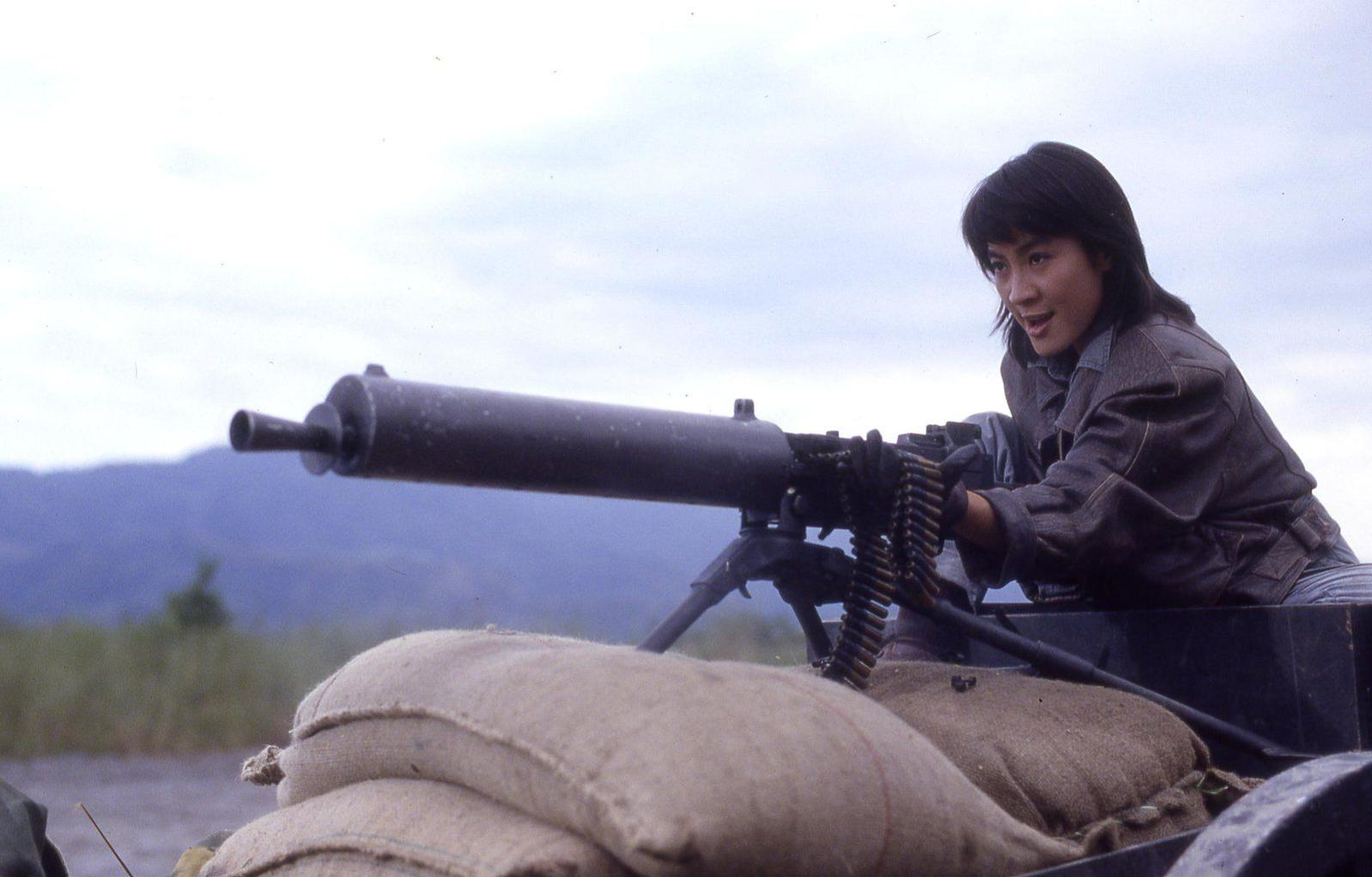 Eine Frau bedient ein Maschinengewehr auf einem Wagen. Sandsäcke bieten Deckung. Berge und ein bewölkter Himmel sind im Hintergrund.