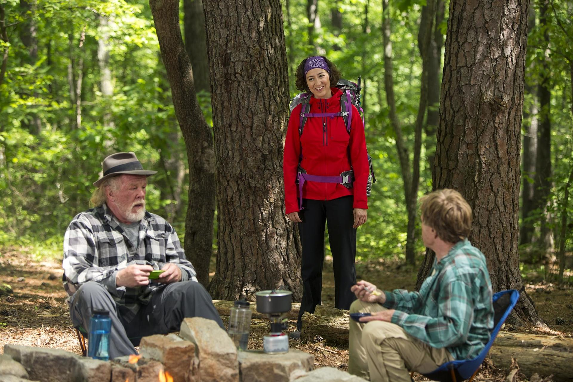 Drei Personen campen in einem Wald. Einer steht, zwei sitzen. Sie befinden sich in der Nähe eines Lagerfeuers, Bäume im Hintergrund.