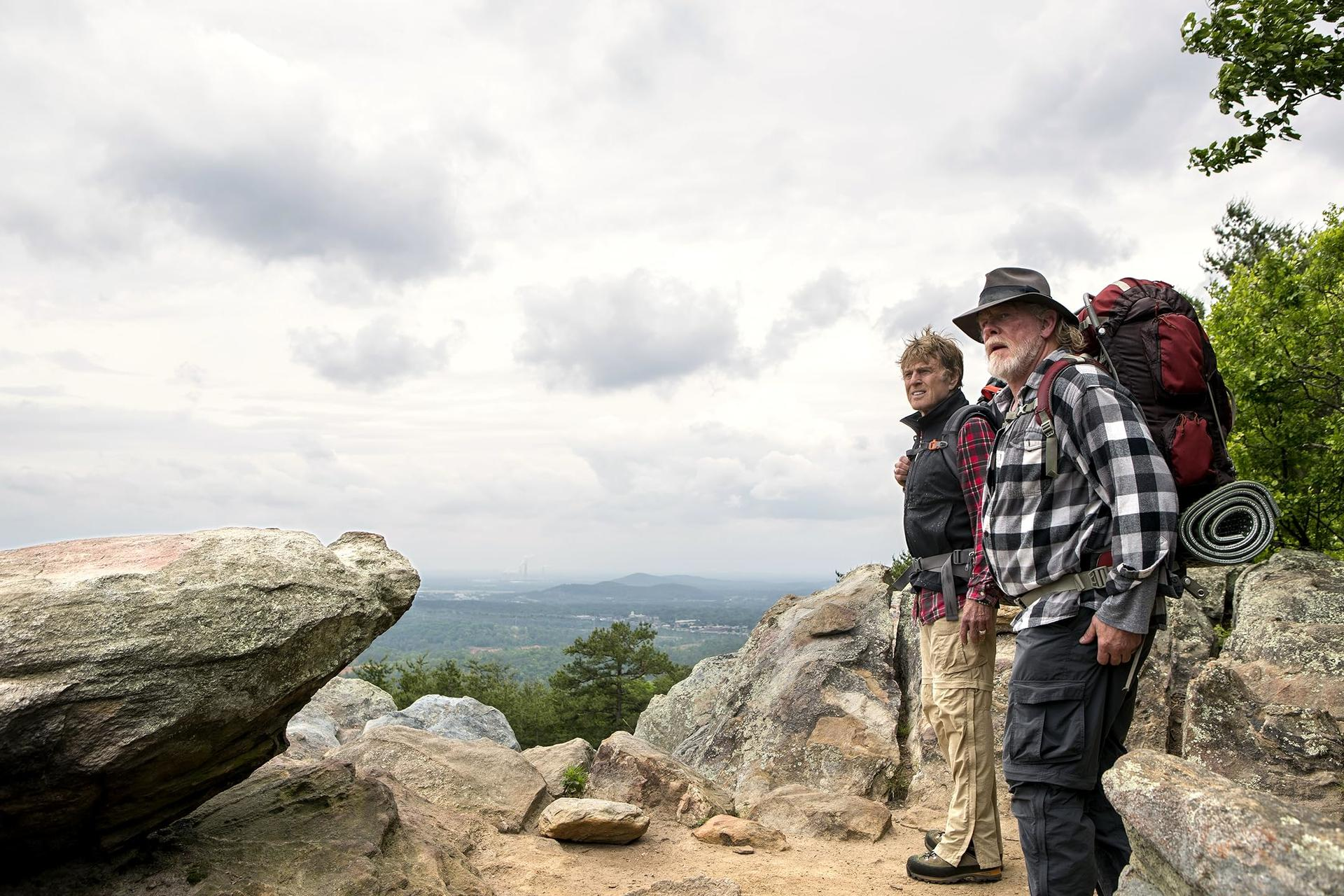 Zwei Männer mit Wanderkleidung auf einem Berg. Wolkenhimmel, Bäume und Tal im Hintergrund. Einer hat einen Rucksack.
