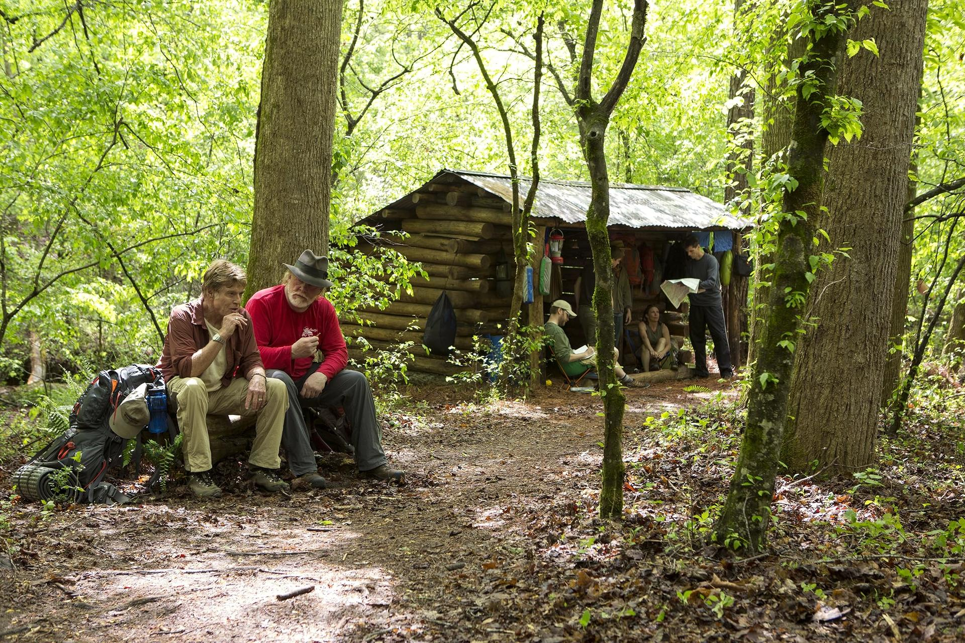 Zwei Männer sitzen in der Nähe einer Hütte in einem Wald. Andere sind in der Nähe. Bäume umgeben die Szene. Rucksäcke liegen am Boden.