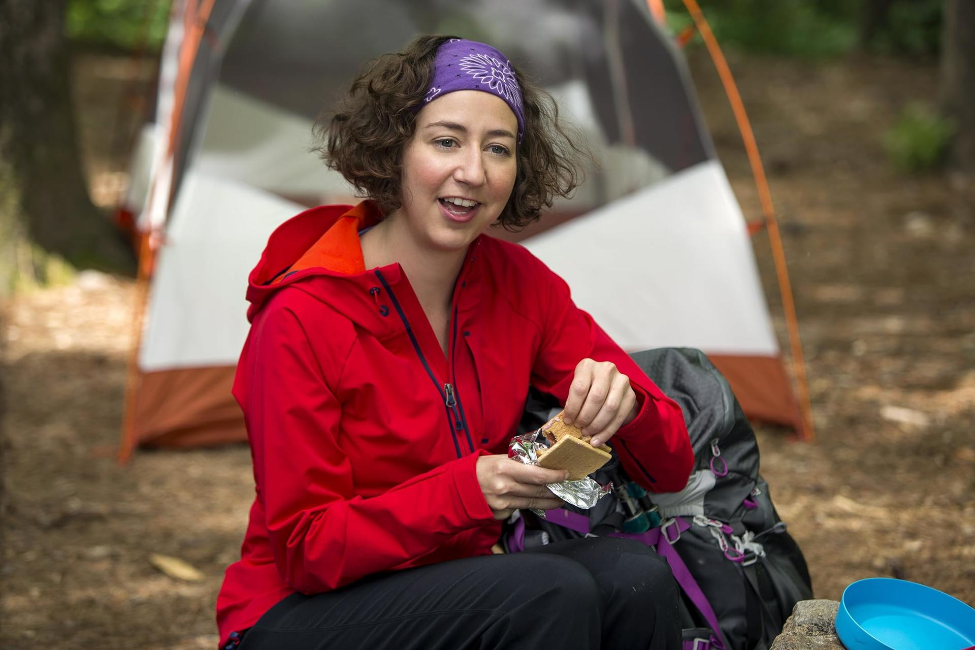 Frau in roter Jacke isst S'more, lila Stirnband, Zelt im Hintergrund. Rucksack und blaue Schüssel davor.