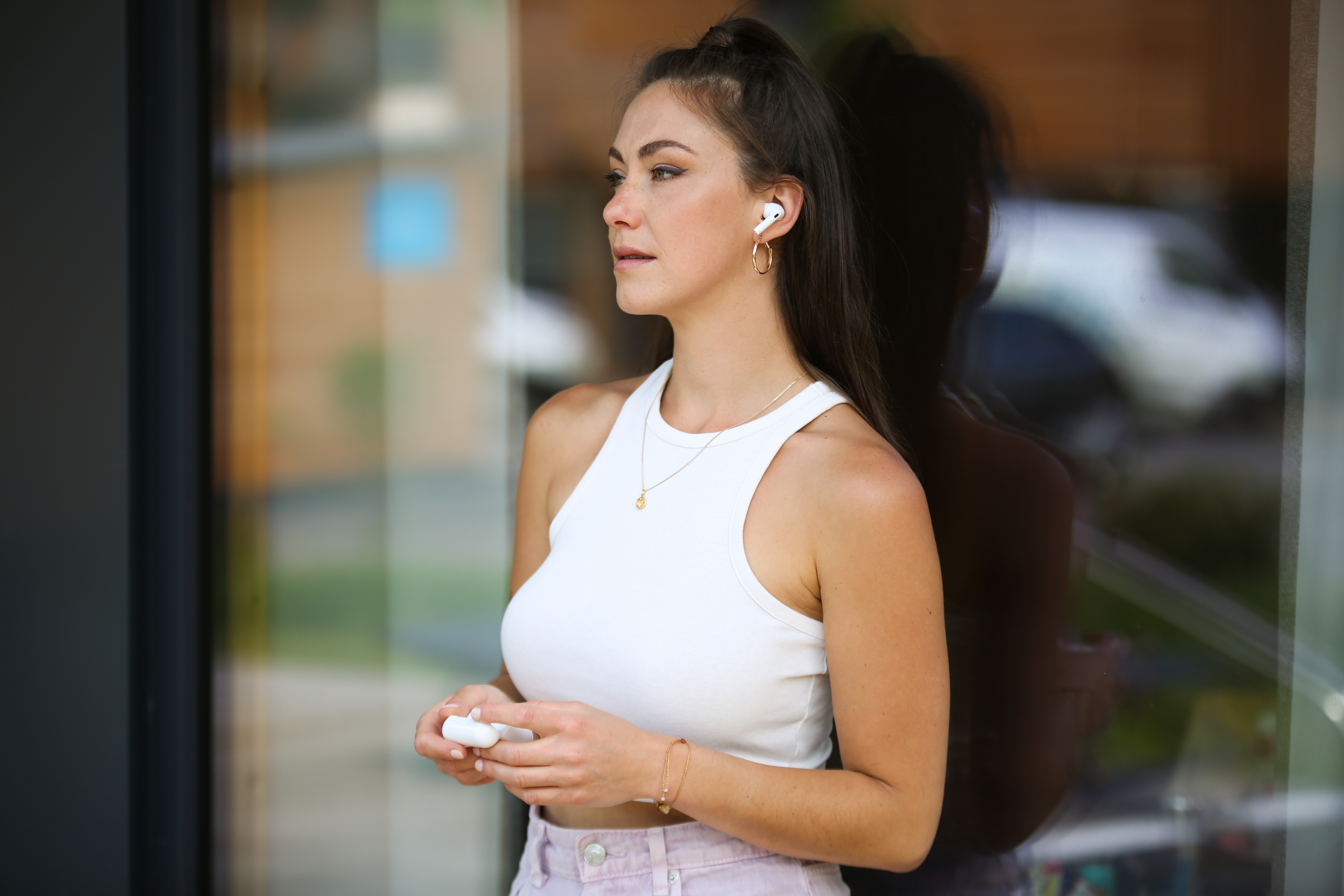 Une femme en débardeur blanc et boucles d'oreilles se tient près d'un mur de verre, tenant des écouteurs. Elle regarde ailleurs, écouteurs en place.