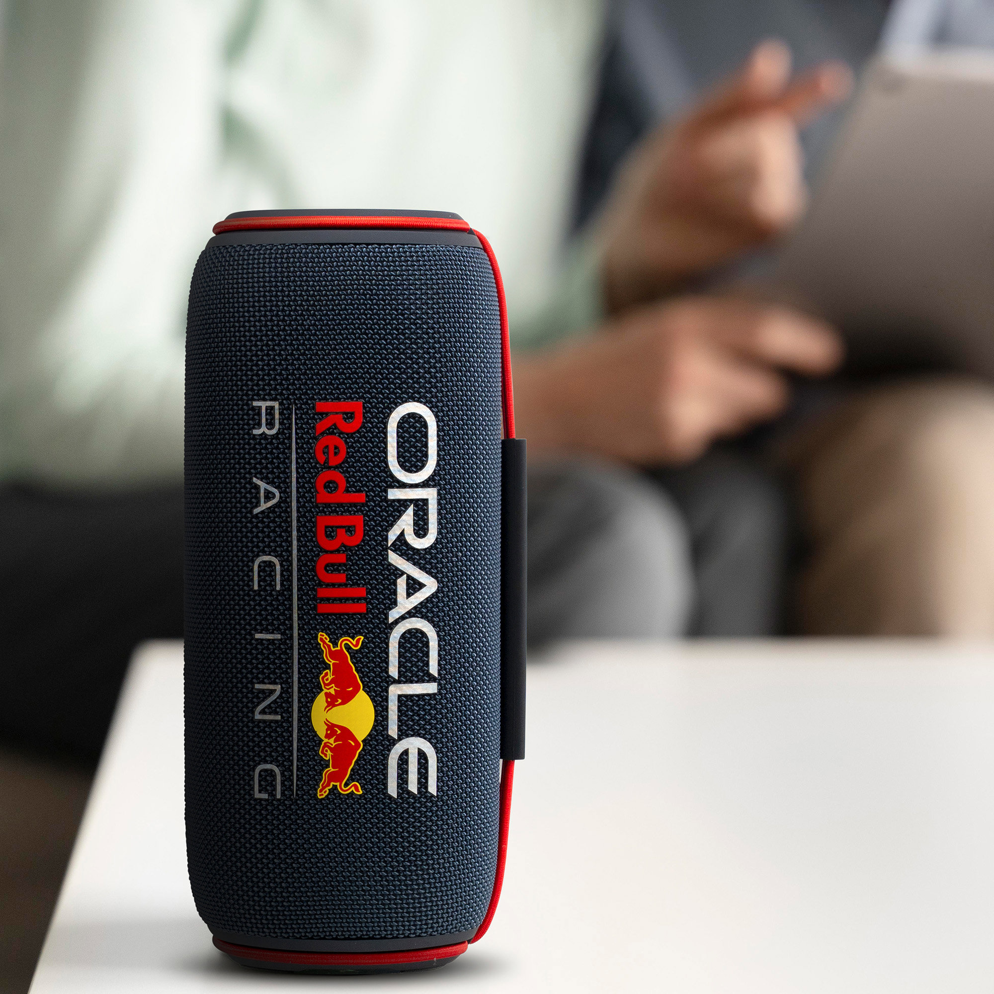 Un haut-parleur Red Bull Oracle est sur une table blanche. Une personne floue en arrière-plan tient une tablette.