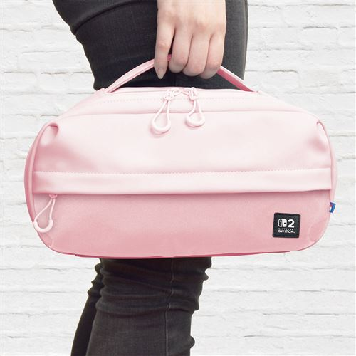 Una persona sosteniendo una funda de transporte rosa para Nintendo Switch. La funda tiene dos cremalleras y un asa. Pared de ladrillo blanco al fondo.