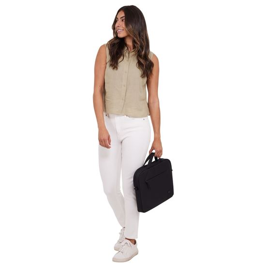 Una mujer con pantalones blancos y top beige lleva una bolsa negra. Mira hacia otro lado, sonriendo, sobre un fondo blanco.