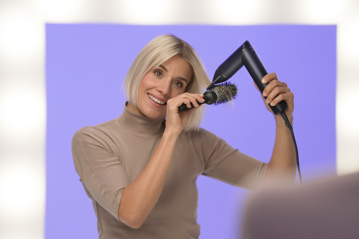 Mujer rubia usando secador y cepillo. Sonríe a la cámara con fondo morado.