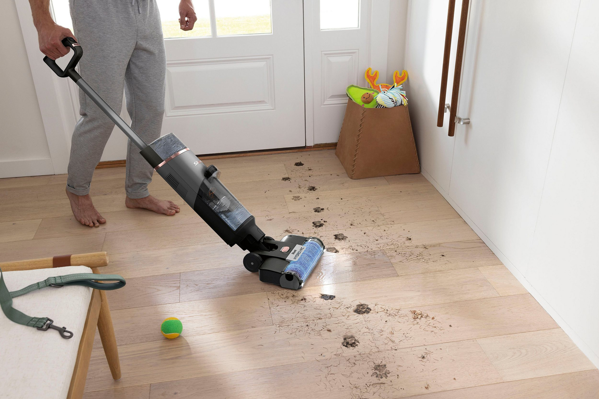 Una persona aspira un suelo de madera sucio. Hay huellas de patas y escombros. Se ven una correa y una pelota para perros. Un cubo con juguetes está en el fondo.
