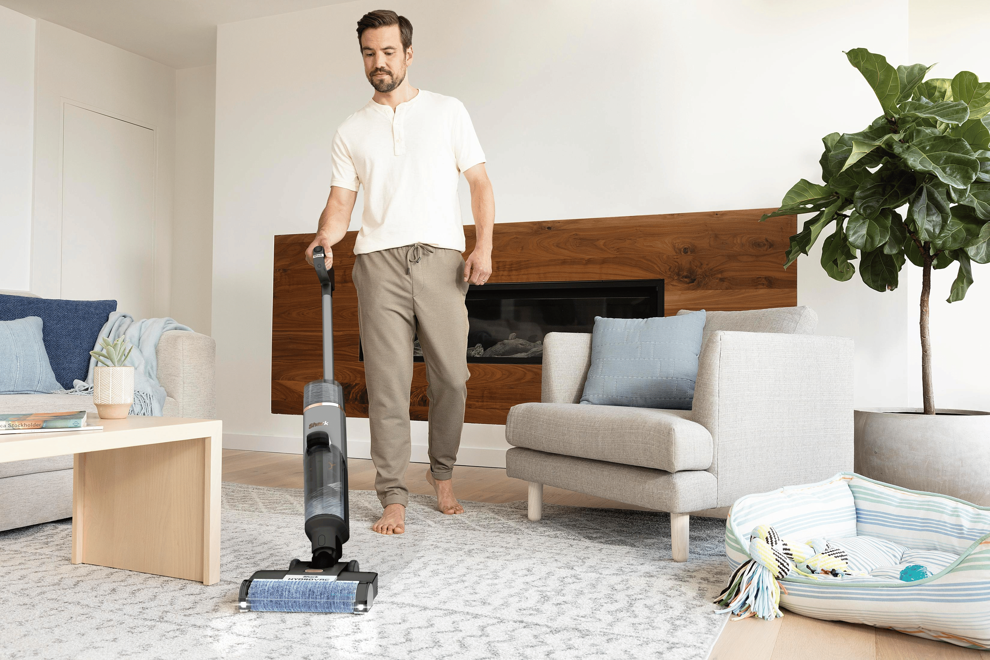Un hombre aspira una sala de estar con una aspiradora moderna. Se ven un sofá, una silla y una planta.