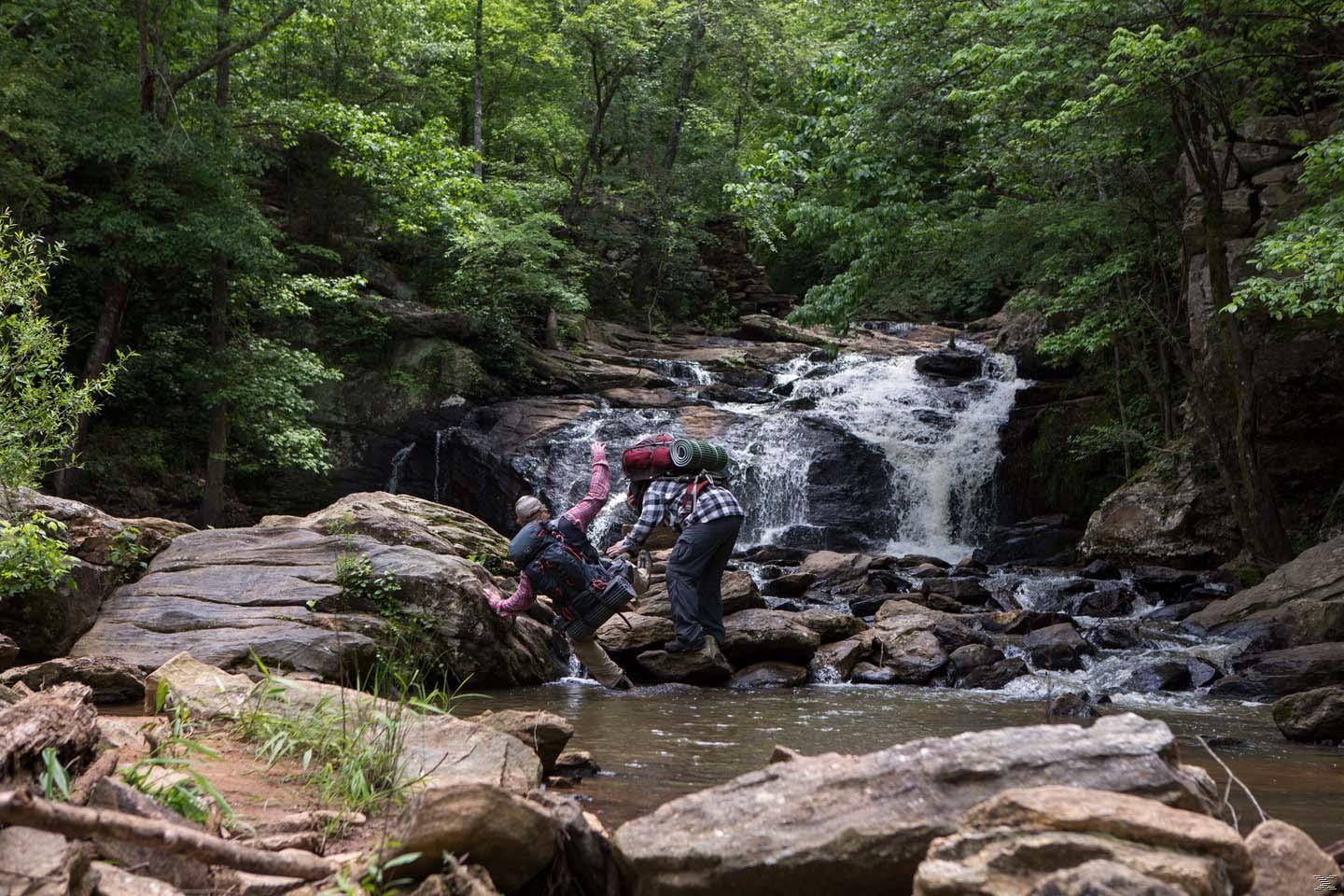 Zwei Wanderer überqueren einen Bach mit einem Wasserfall im Hintergrund, umgeben von Bäumen und Felsen.