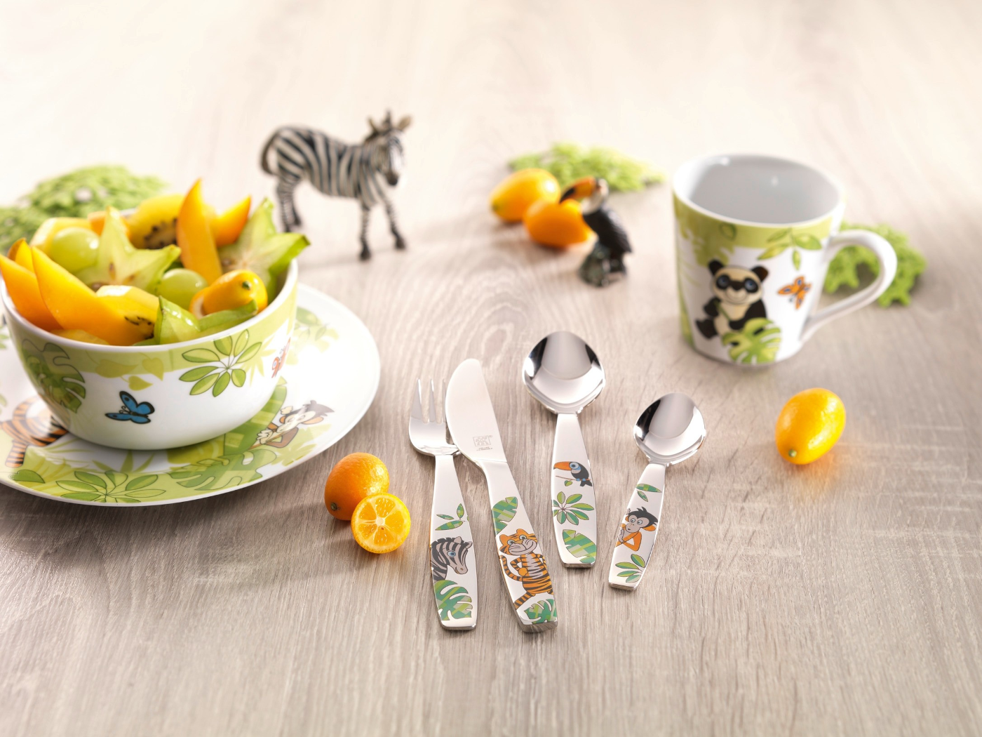 Eine Schüssel mit Obst, Besteck mit Tierdesigns, eine Tasse mit Panda und Spielzeugtiere auf einer Holzoberfläche.