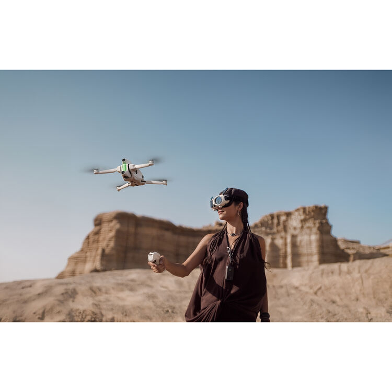 Een vrouw met een bril bestuurt een drone met een afstandsbediening. De drone vliegt boven een woestijnlandschap met rotsformaties en een blauwe lucht.
