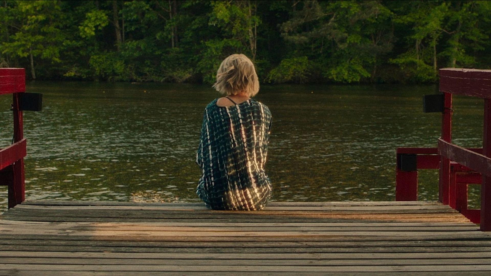 Eine Frau sitzt auf einem Holzsteg mit Blick auf das Wasser. Bäume säumen das Ufer. Rote Stegstützen rahmen die Sicht.
