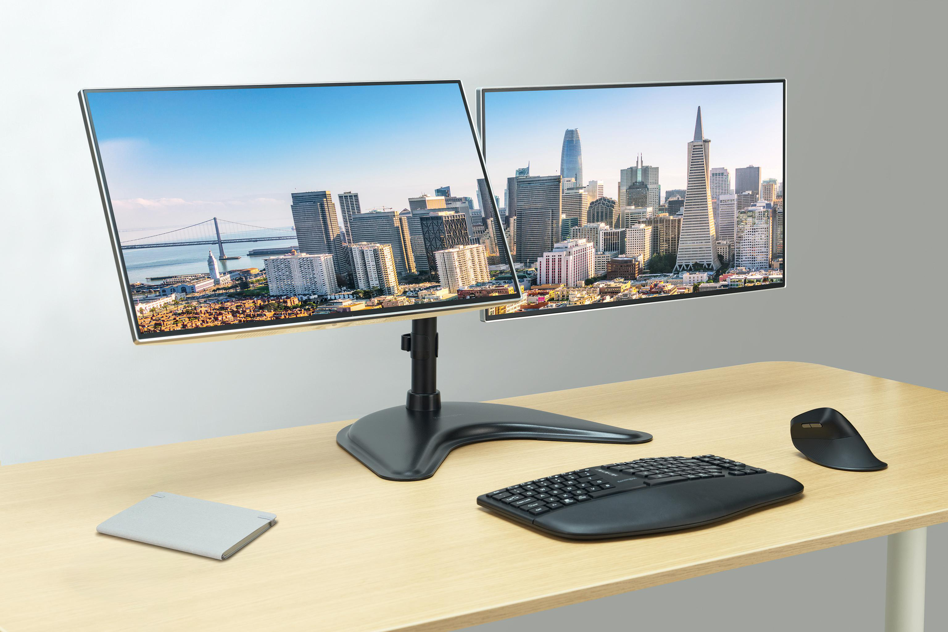 Zwei Monitore zeigen eine Skyline. Tastatur, Maus und Notizblock liegen auf einem Holztisch.