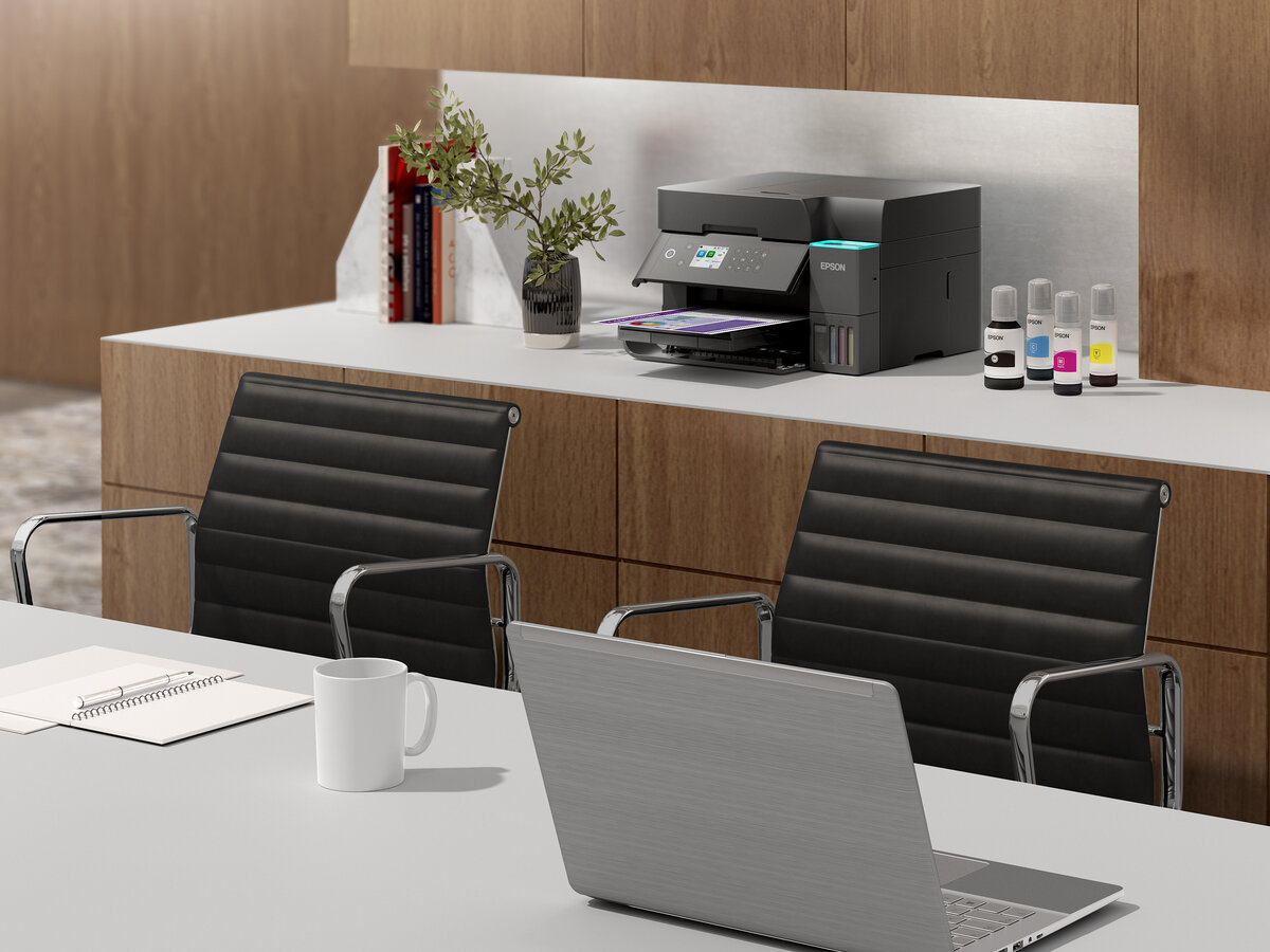 Scena biurowa z drukarką, butelkami z tuszem i laptopem. Czarne krzesła, kubek i notatnik również znajdują się na biurku.