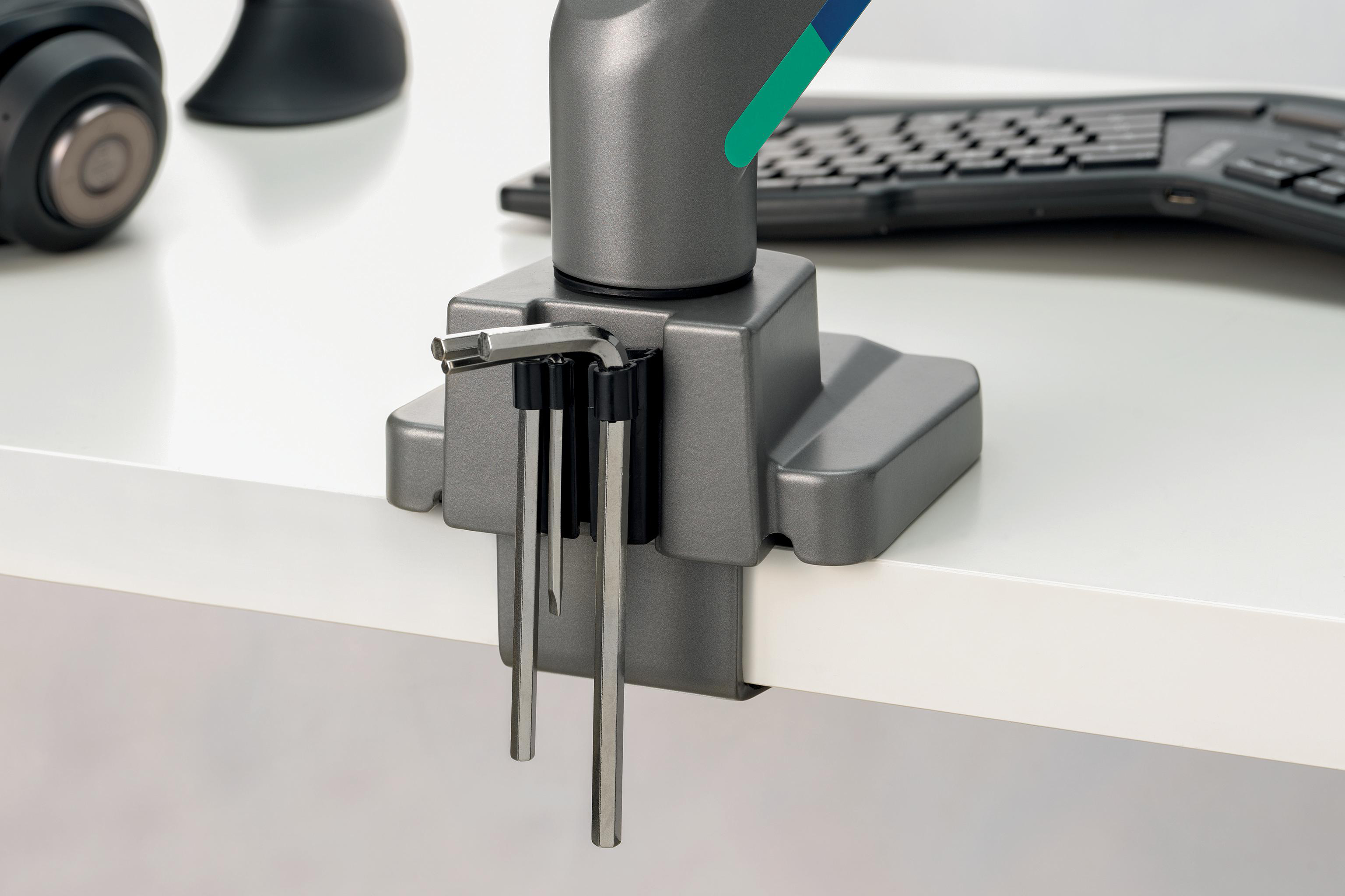Graue Schreibtischklemme mit Inbusschlüsseln. Tastatur und Headset sind im Hintergrund auf einem weißen Schreibtisch zu sehen.
