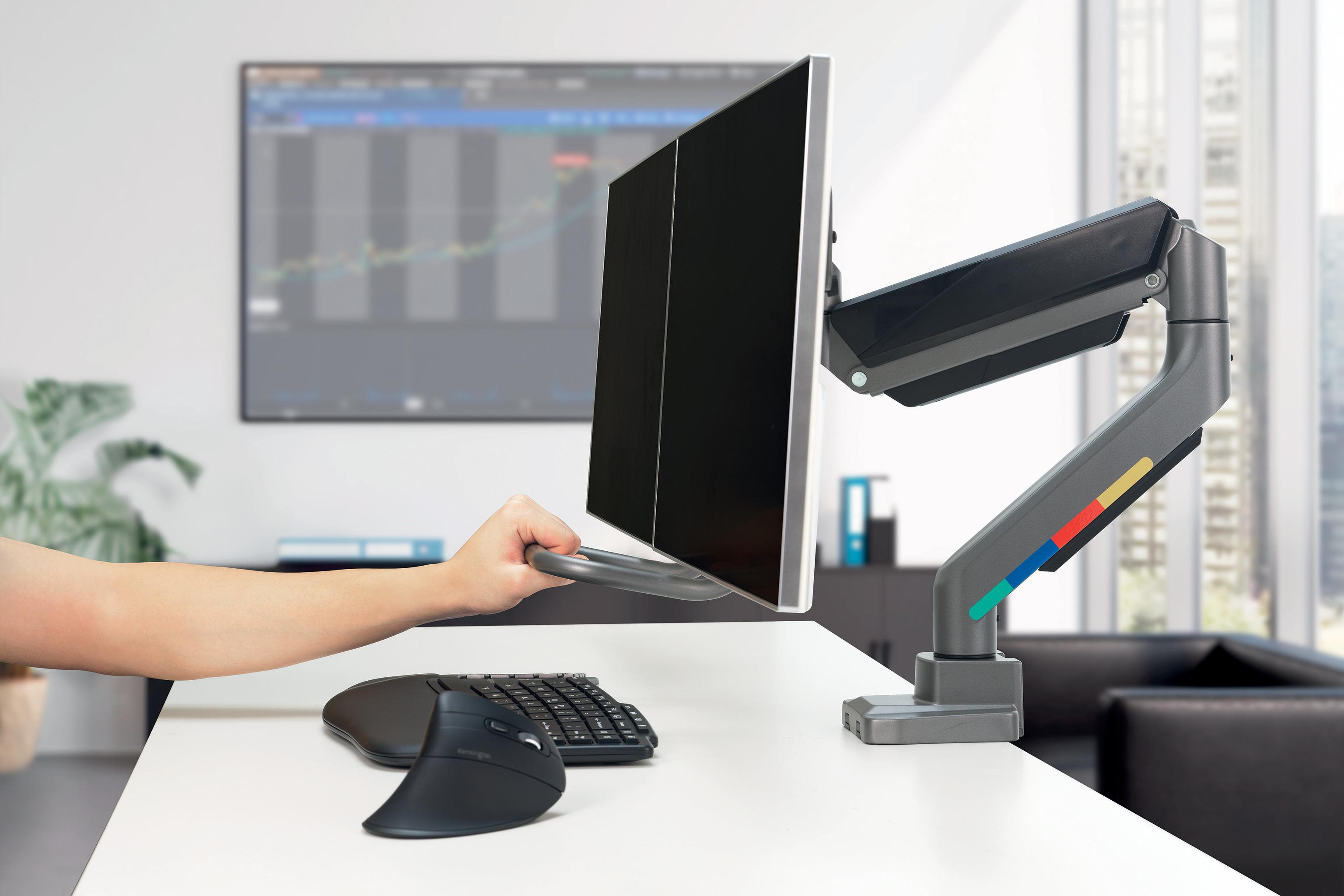 Eine Hand justiert einen Monitor auf einem Schreibtisch. Tastatur und Maus sind vorhanden. Ein Diagramm ist auf dem Hintergrundbildschirm.