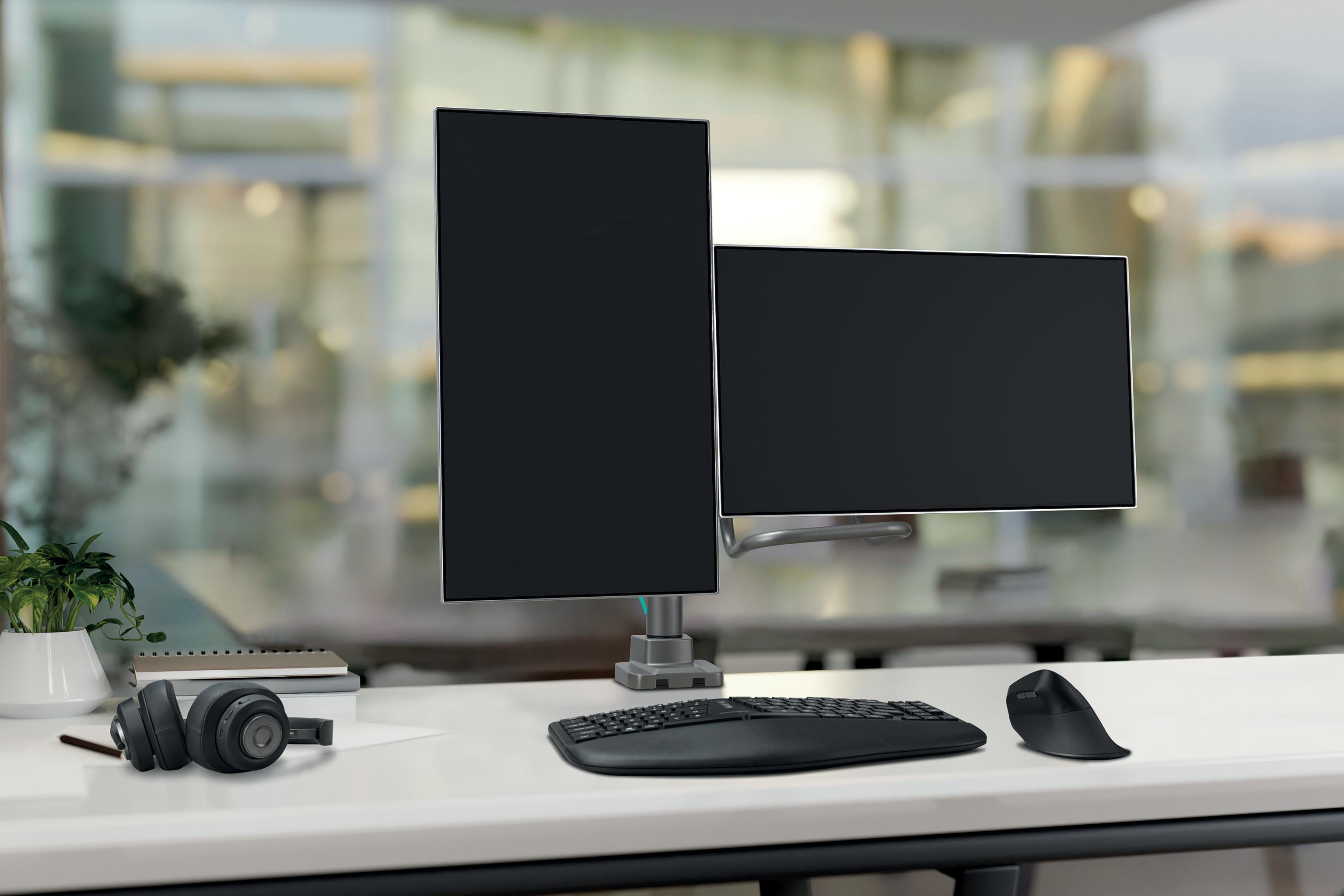 Schreibtisch mit zwei schwarzen Monitoren, Tastatur, Maus, Kopfhörern und einer Pflanze. Verschwommener Bürohintergrund.
