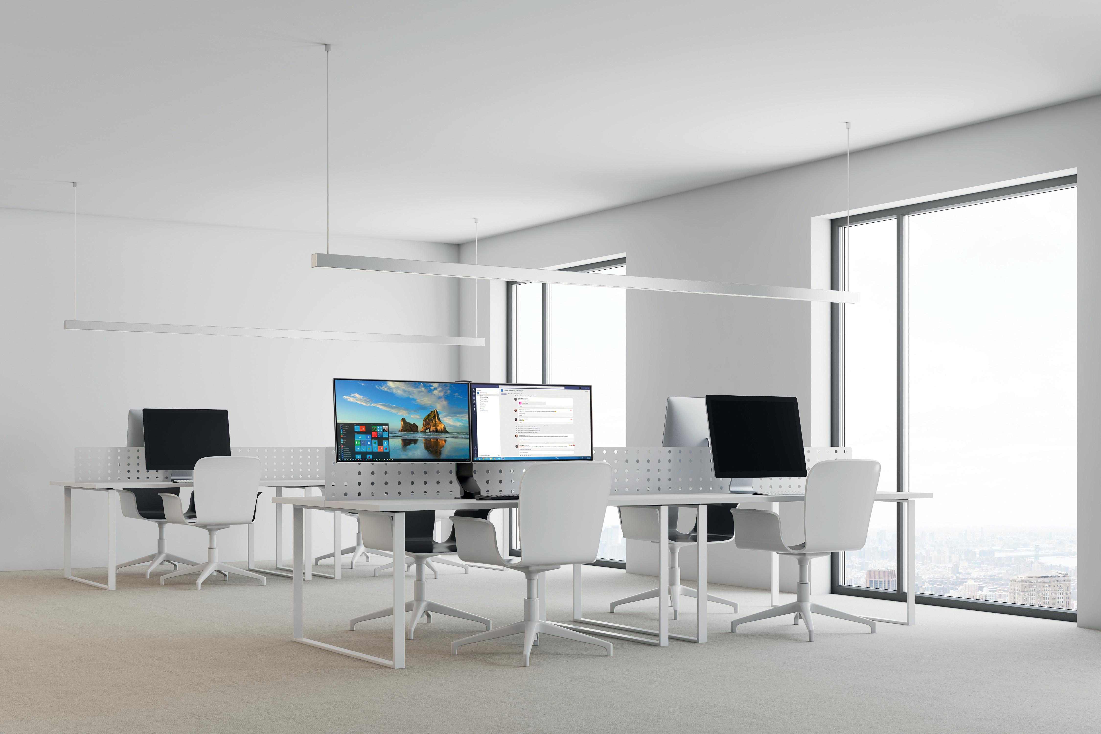 Büro mit Schreibtischen, Computern und Stühlen. Große Fenster zeigen eine Stadtlandschaft. Der Raum hat weiße Wände und einen hellen Teppich.