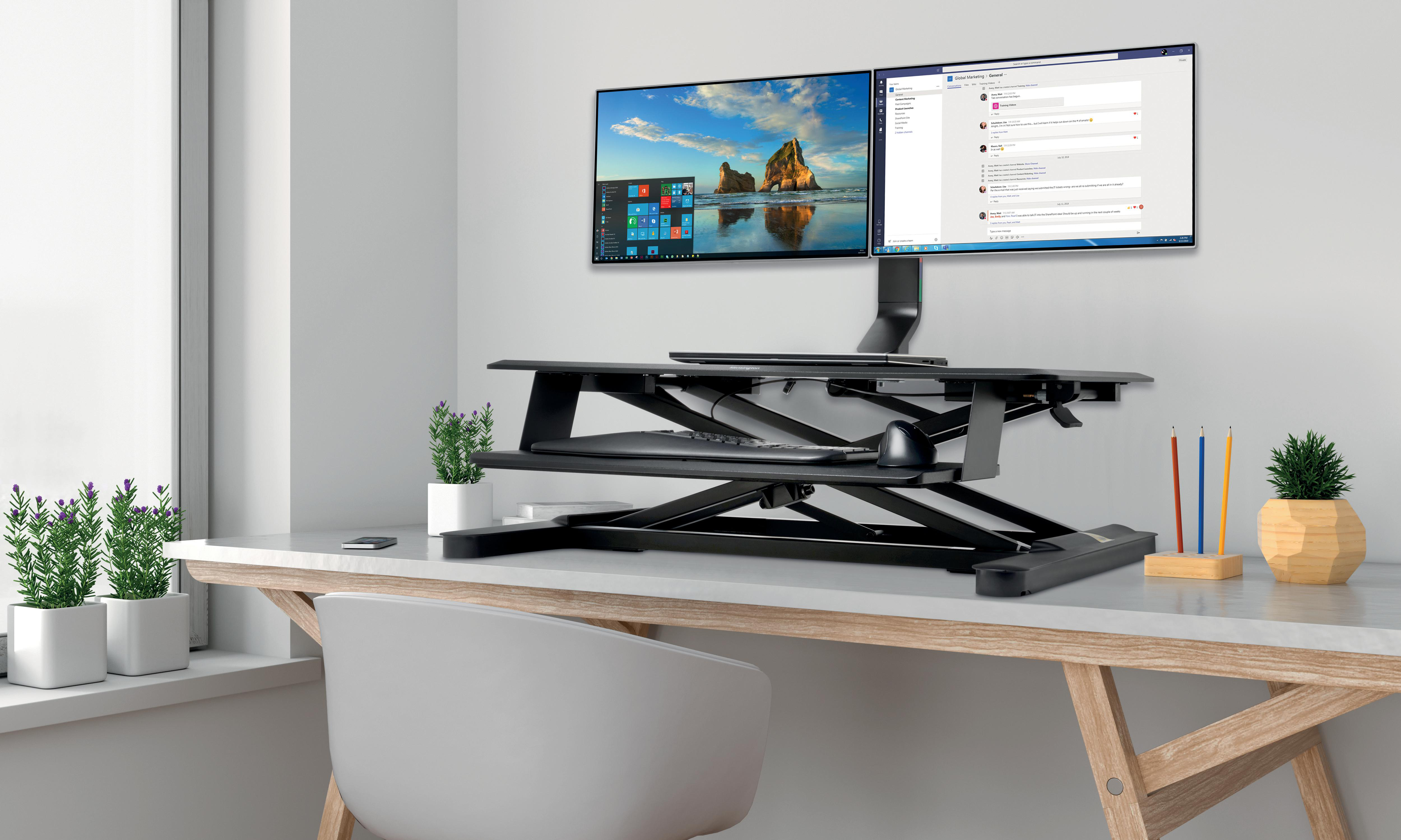 Ein Schreibtisch-Setup mit zwei Monitoren, einem schwarzen Ständer, einer Tastatur, einer Maus, Pflanzen und einem Stuhl. Links ist ein Fenster.