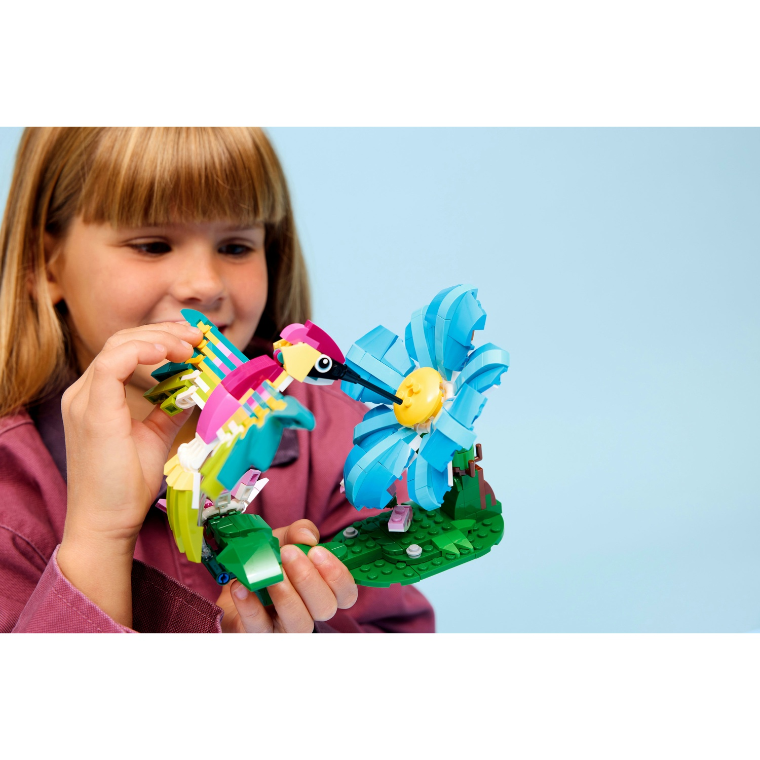 Una ragazza tiene in mano un uccello Lego e un fiore blu. L'uccello è colorato, il fiore blu con centro giallo. Sfondo azzurro.