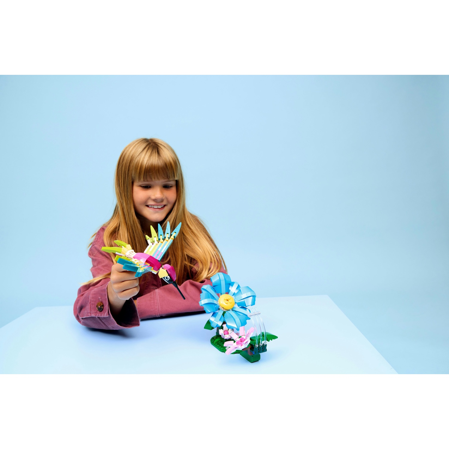 Una ragazza tiene in mano un colorato colibrì Lego. Un fiore Lego è sul tavolo. Lo sfondo è azzurro.